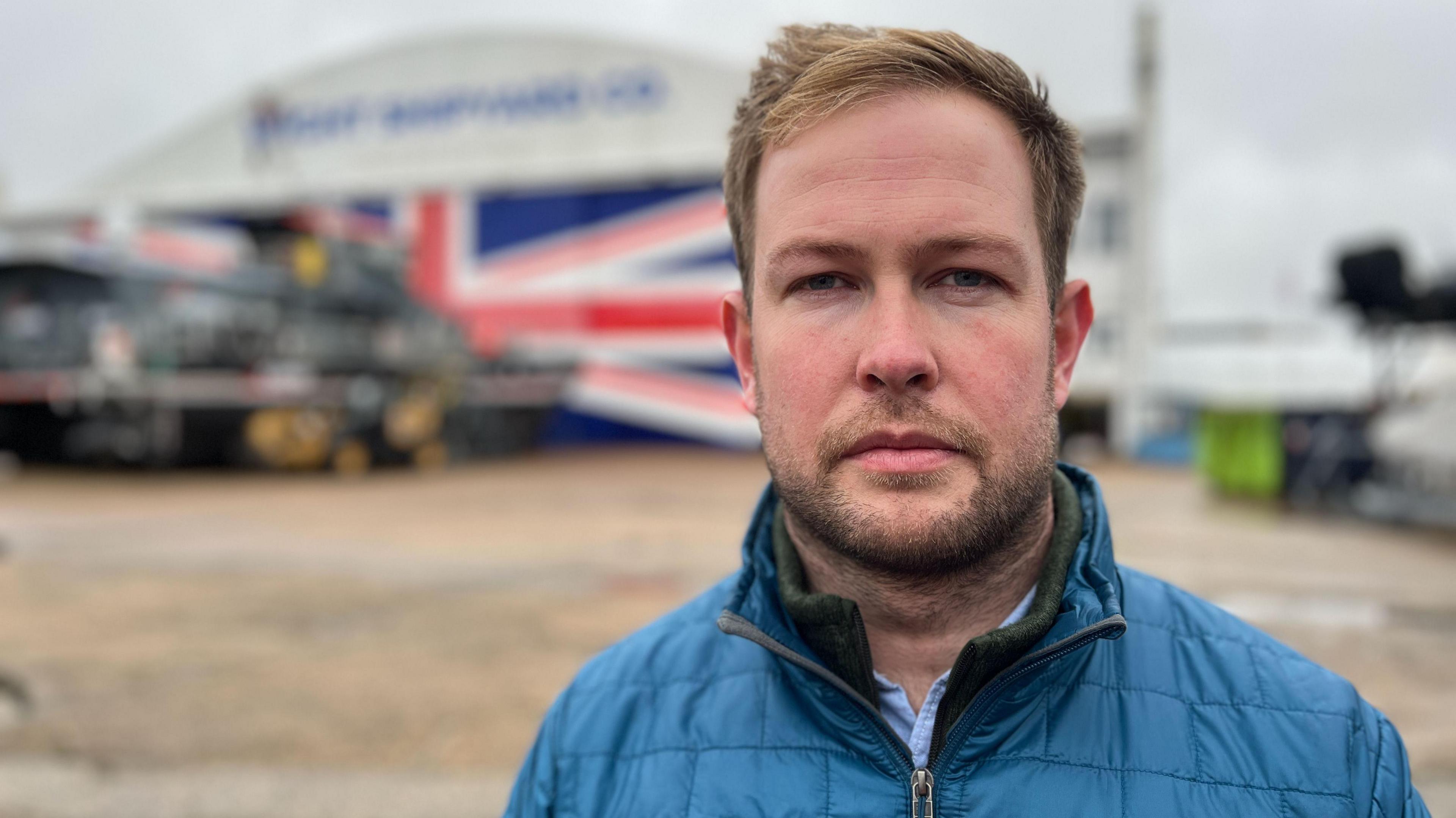 Toby Mumford, has short auburn hair and brown stubble. He is wearing a blue coat. Behind him, out of focus is a large airport hangar-type structure.