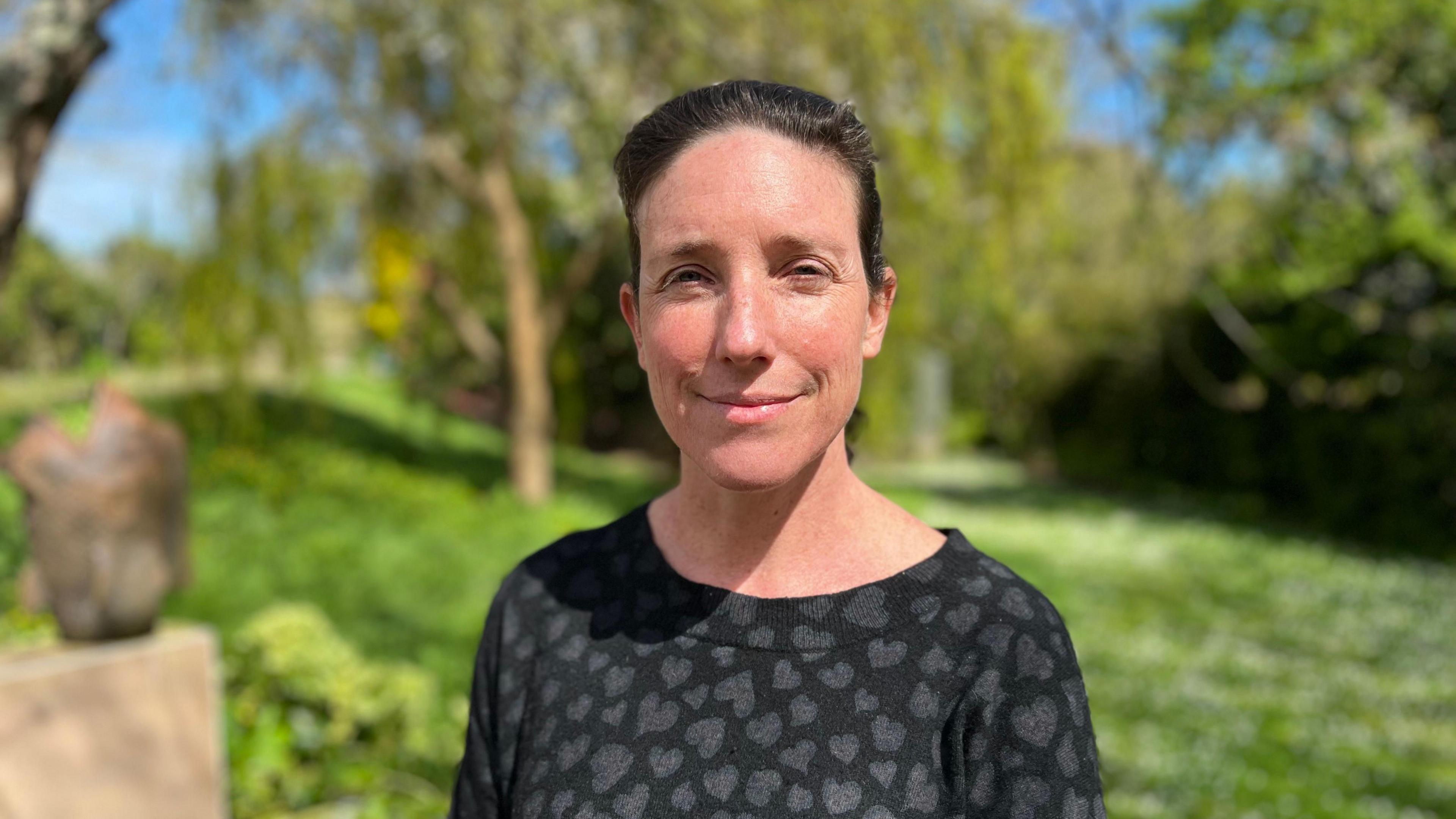 President of the Committee for Environment and Infrastructure, Deputy Lindsay De Sausmarez stood in green garden, with trees in the background. She is in a black top and she is slightly squinting in the sunlight. 