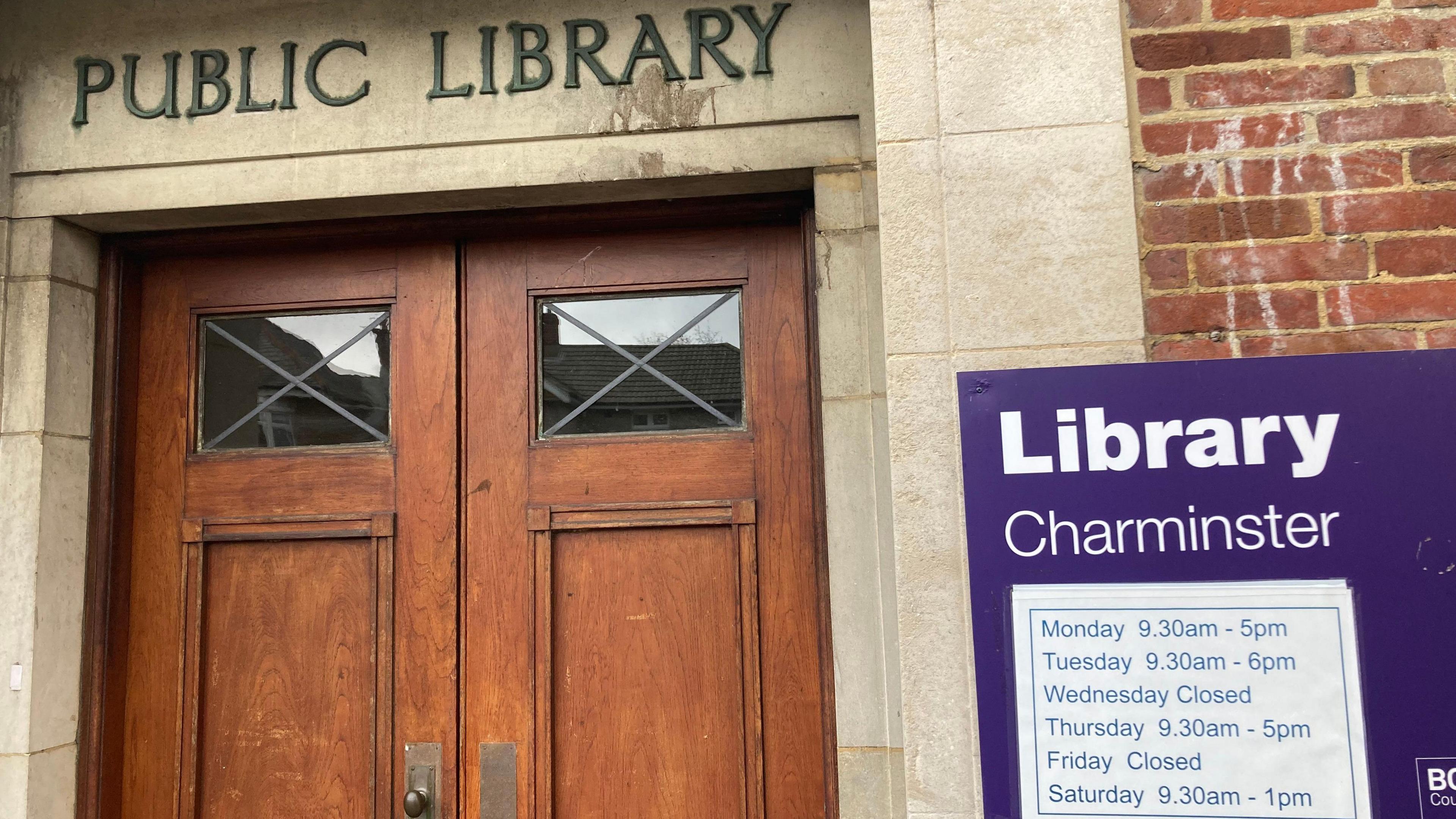 A solid wooden door at Charminster library. A sign with library opening hours is on the right of the picture