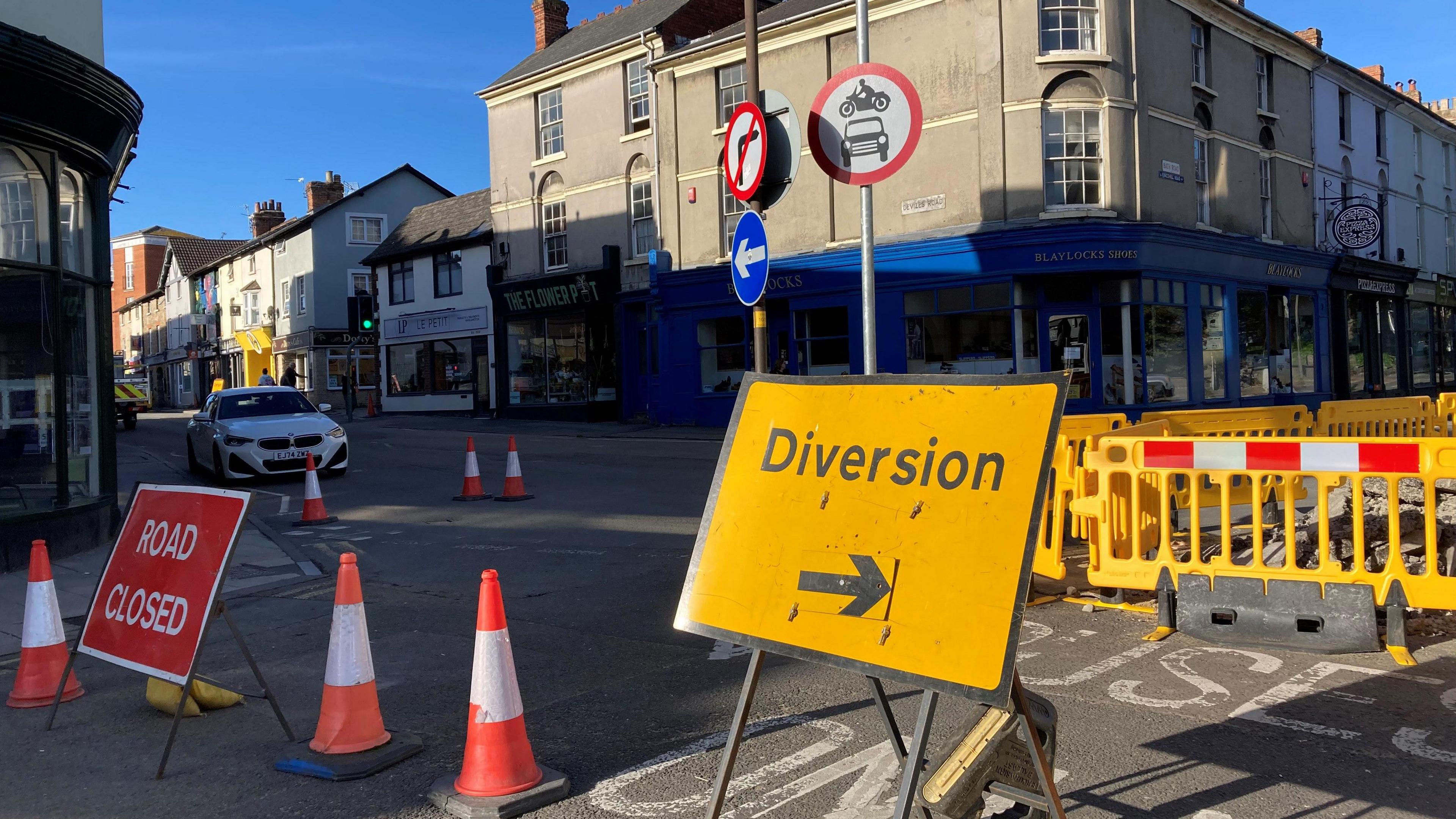 Several road signs saying 'Road Closed and Diversions, in front of road junction near shops and flats.