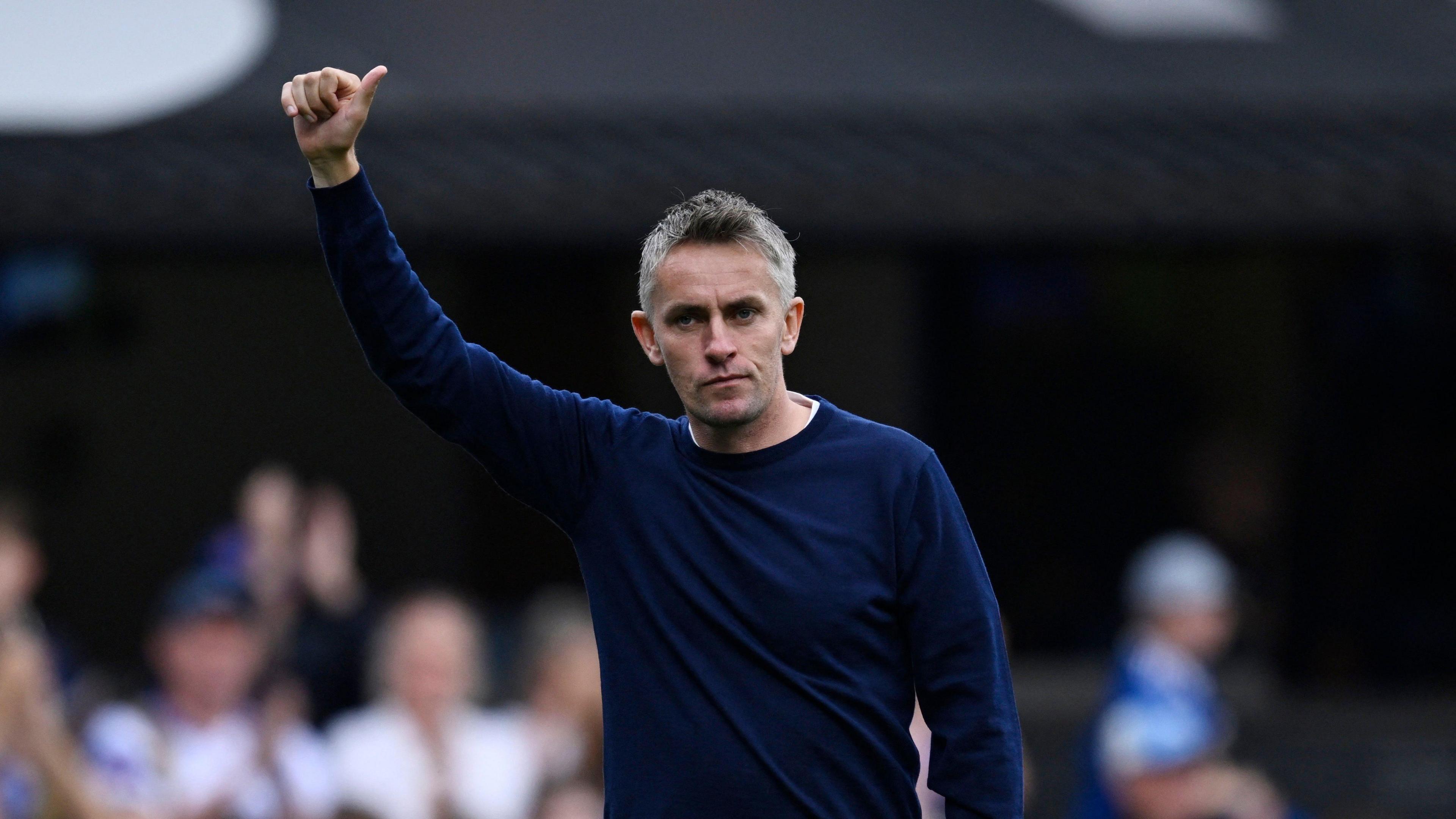 Ipswich Town manager Kieran McKenna. He has one hand up in the air making a thumbs up gesture to supporters. He is wearing a navy jumper.