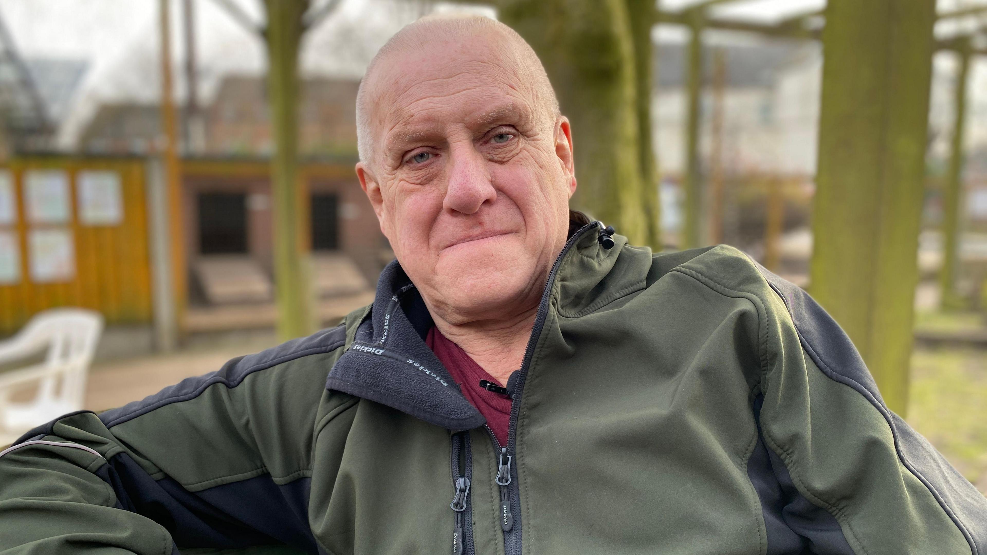 Tony Coles is wearing a green long-sleeved jacket and a red top. He is sitting on a bench in the farm with wooden huts in the background.