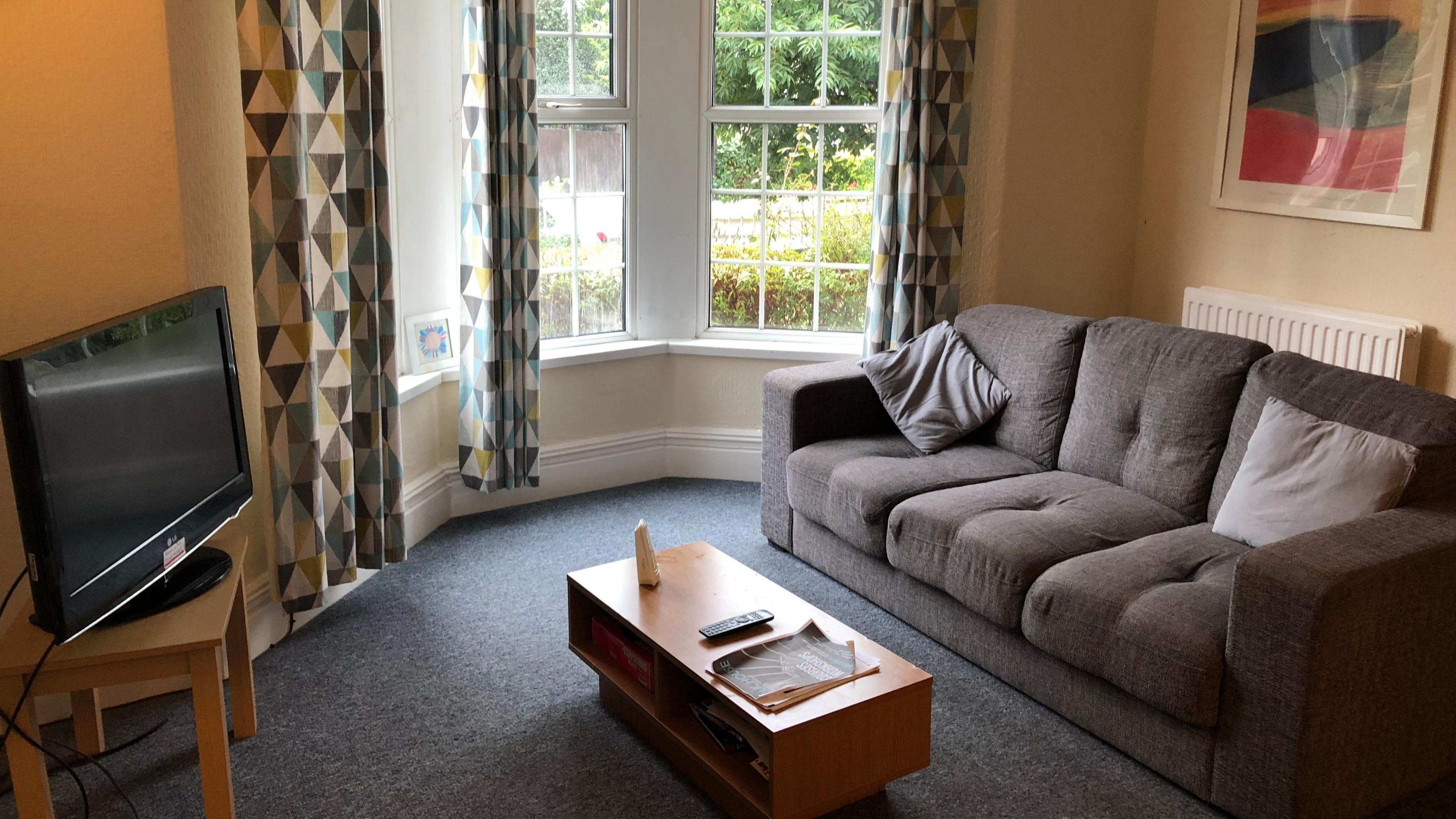 A living room, which will be tenanted by someone on the scheme. There is a tv stand in the corner of the room, a large and bright bay window with long, geometric patterned curtains. There is a wooden coffee table to the centre and a grey three-seater sofa with cushions. An abstract painting is being hung on the wall. The room is magnolia in colour with grey carpeted flooring.