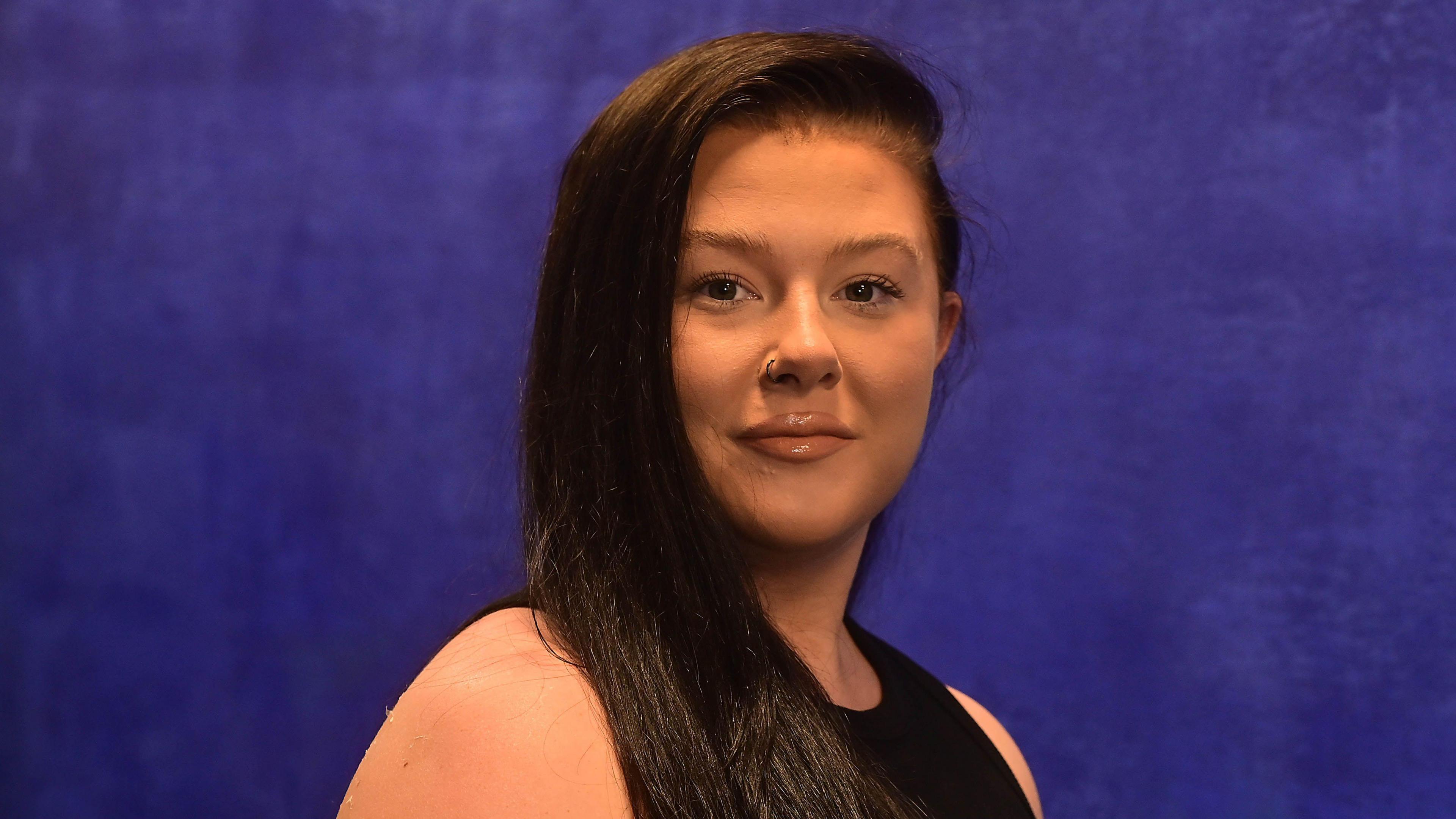 Ms Storey is standing in front of a blue backdrop. She has long brown hair and is wearing a black top and has a hoop piercing on her nose. 