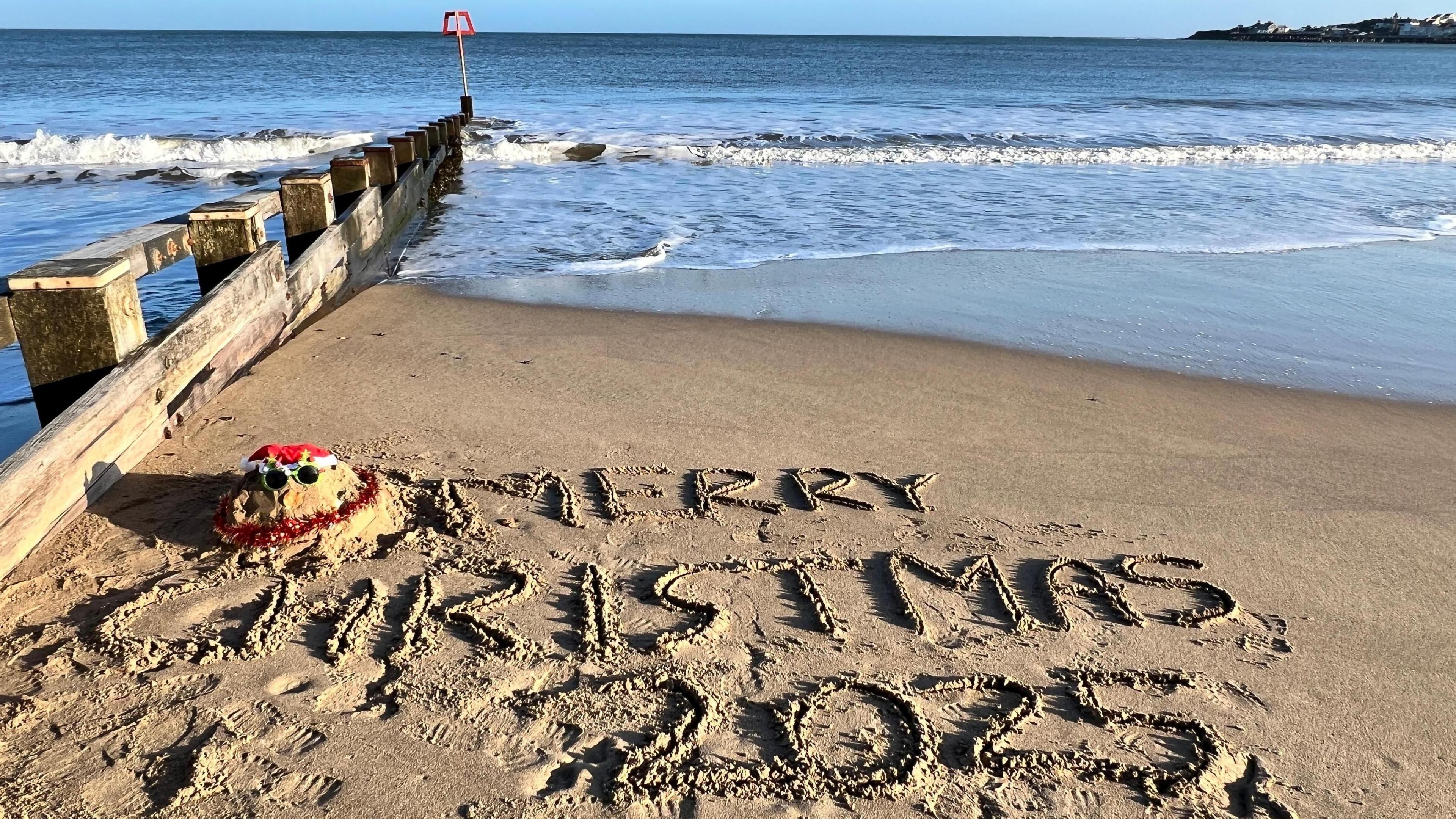 Swanage, Dorset, BBC Weather Watcher, Don Don snapped a photo of Merry Christmas 2025 written in the sand at Swanage with waves lapping and a snad snowman with santa hat, tinsel and festive glasses