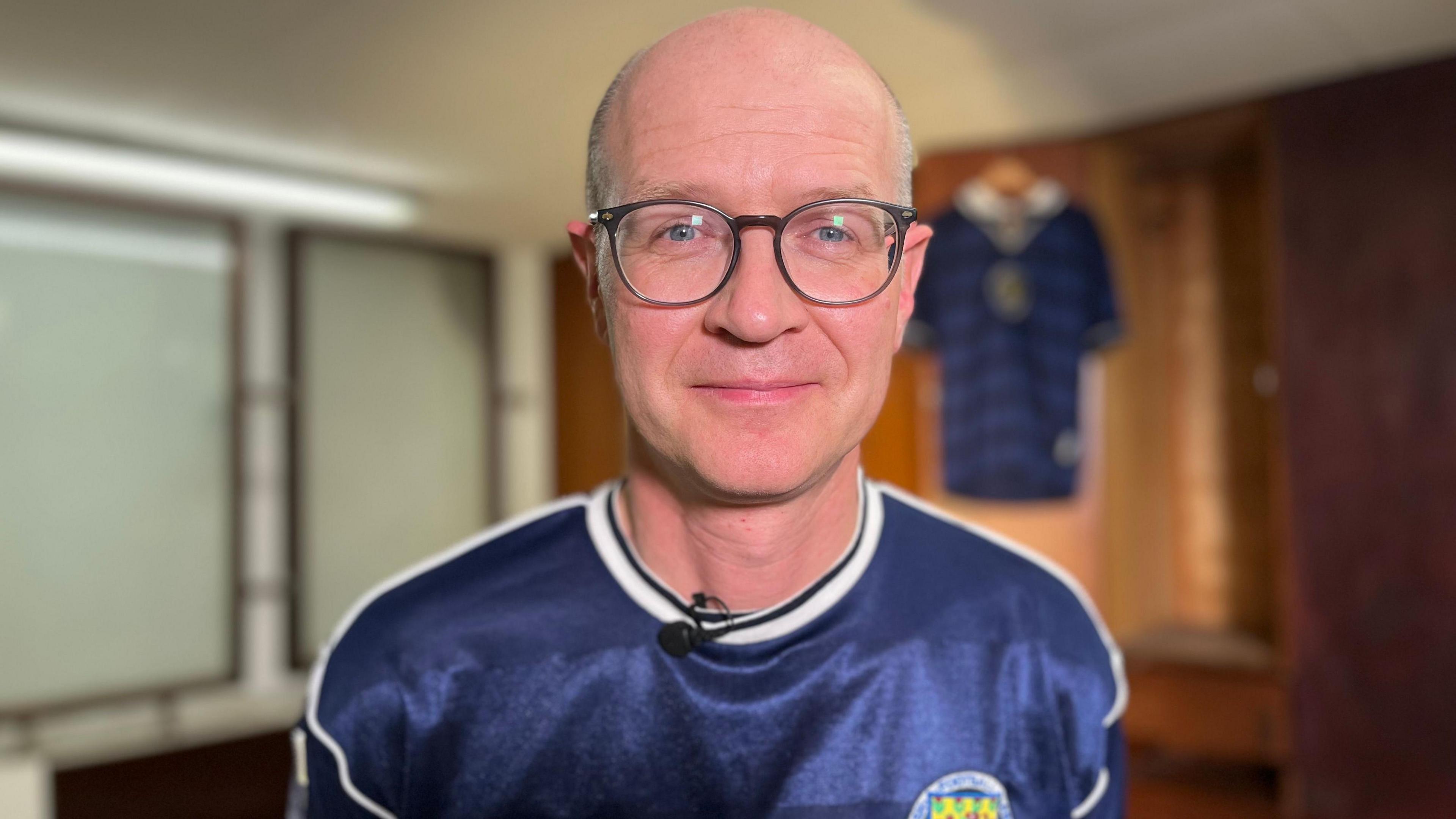 Henry Hepburn, a bald man with glasses wearing a Scotland jersey smiles at the National Football museum, a Scotland jersey handing on the wall behind him.