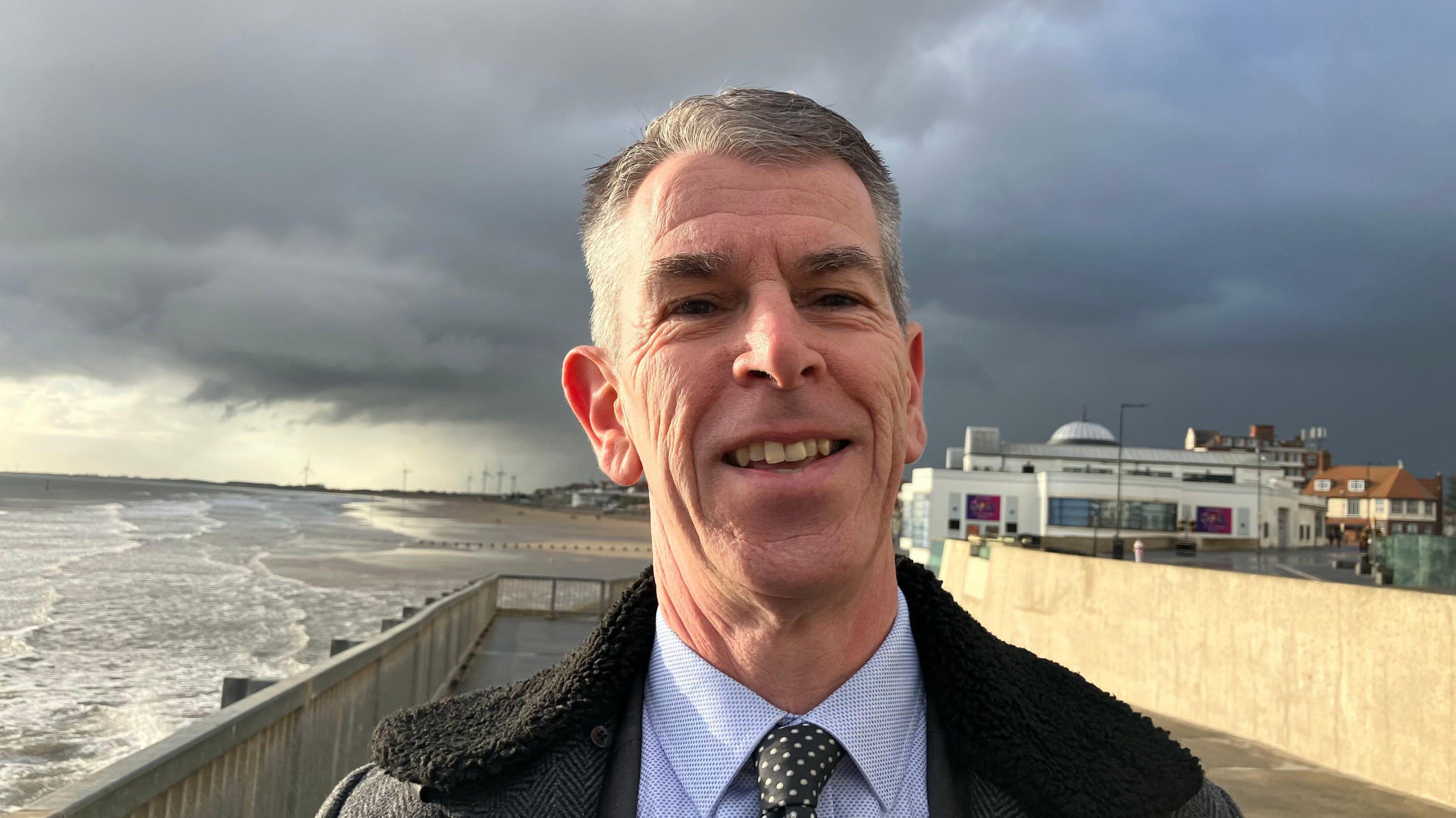 A man with short grey hair is smiling and wearing a black coat, blue shirt and black and white spotty tie. He is standing on a promenade next to a beach. The sky is cloudy.