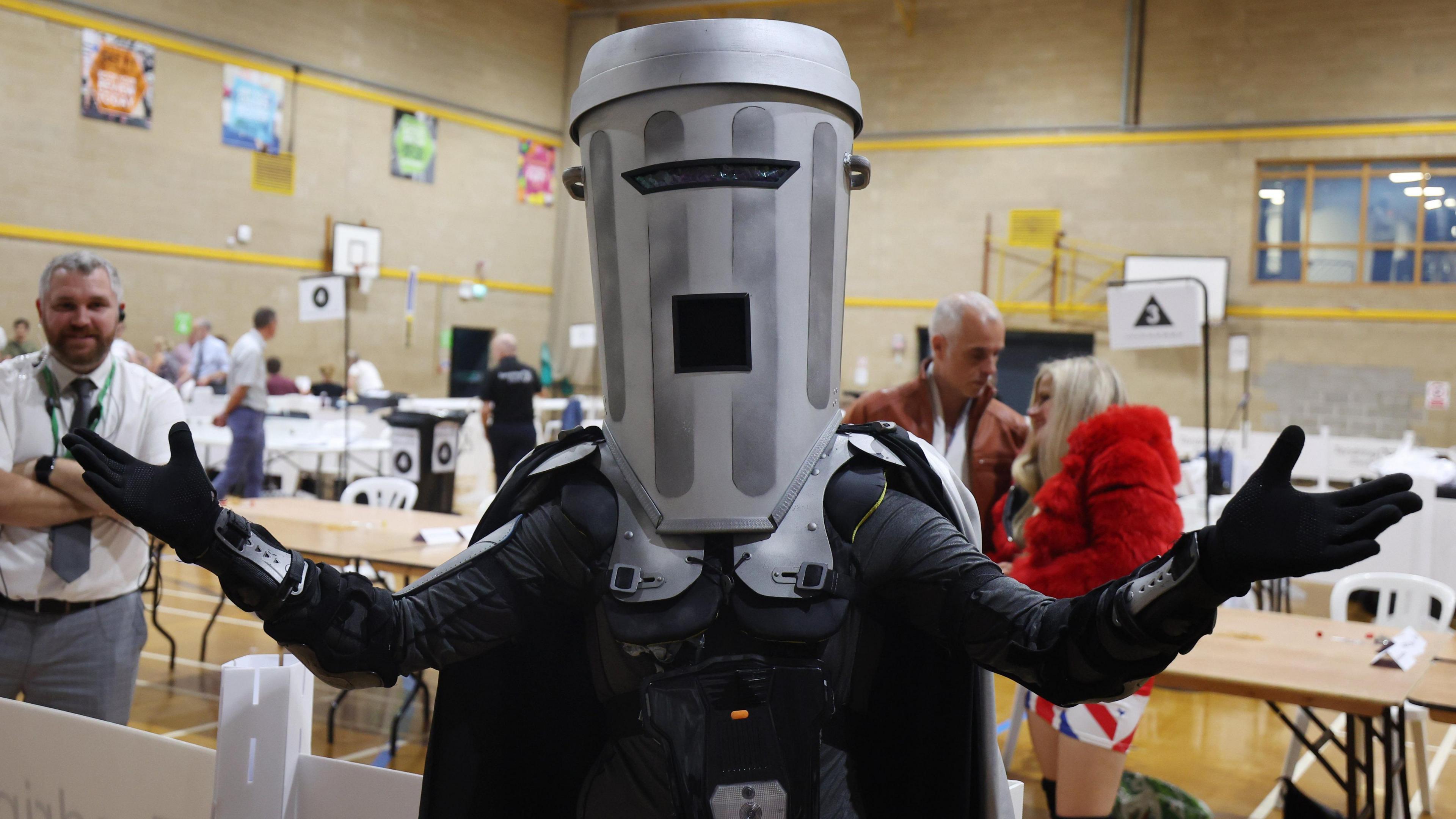 A person wearing an elaborate sci-fi costume with a large cylindrical helmet and dark armour stands with arms outstretched inside a sports hall, while attendees and display tables are visible in the background.
