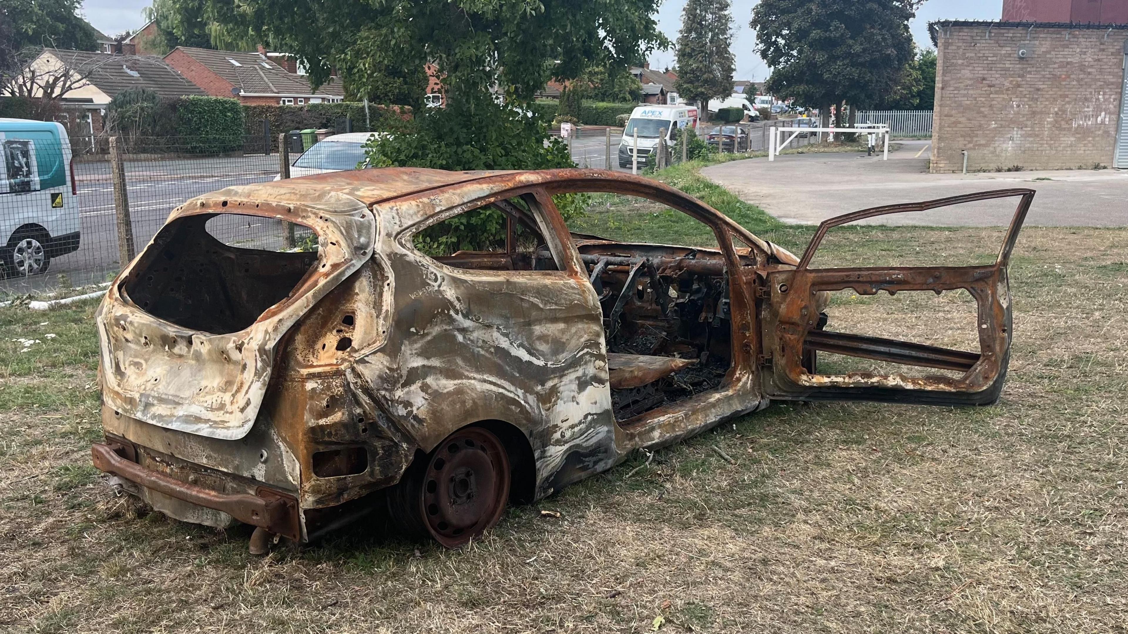 A burnt out car in a park. There is a building on the right hand side and a road with cars and vans on it in the background.