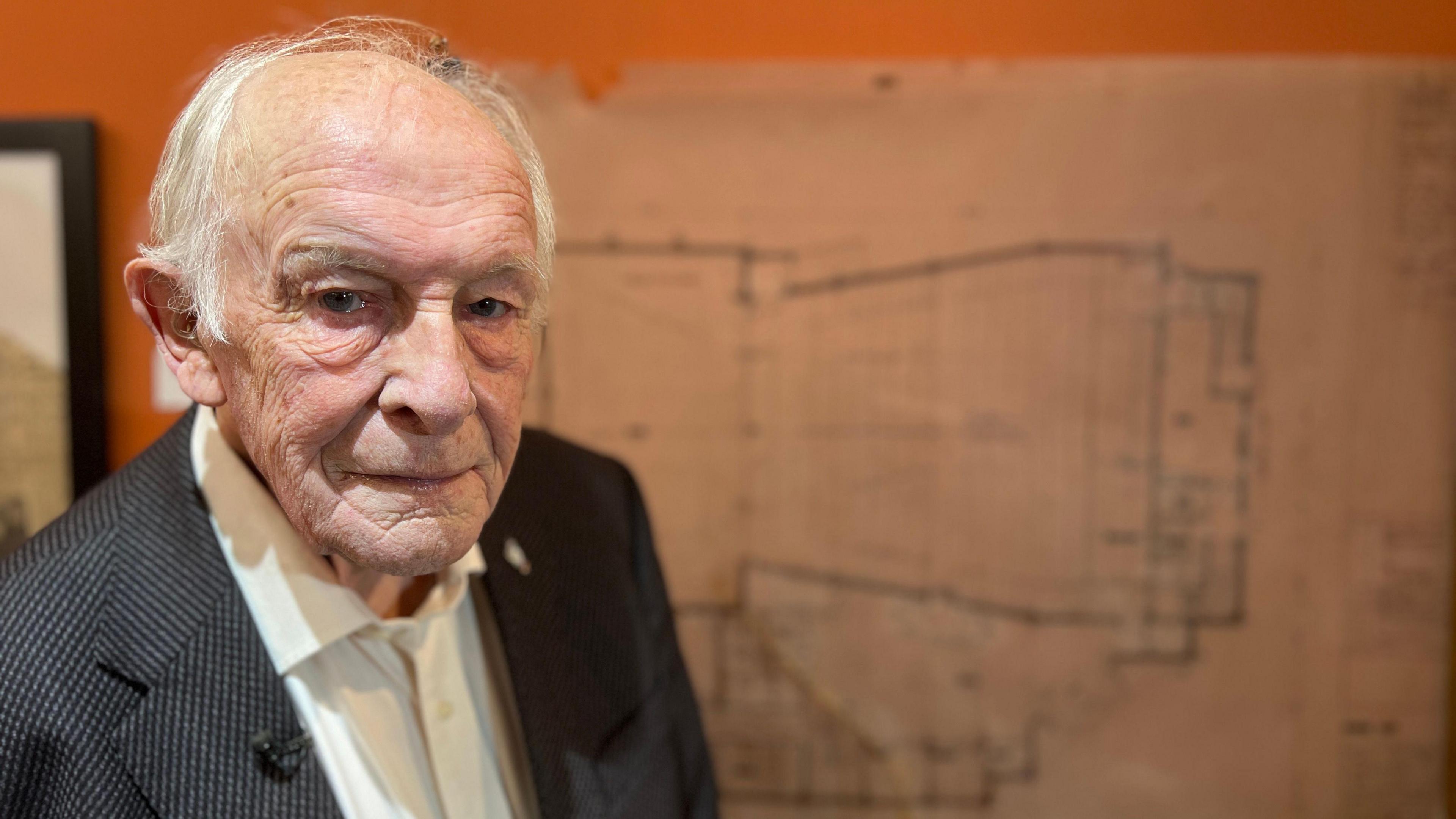 An old man with thin grey hair, who is wearing a white shirt and a black suit jacket, looks at the camera as he stands in front of an orange wall where his browned architectural plans for The Roses Theatre, which he drew in the 1970s, are on exhibition.
