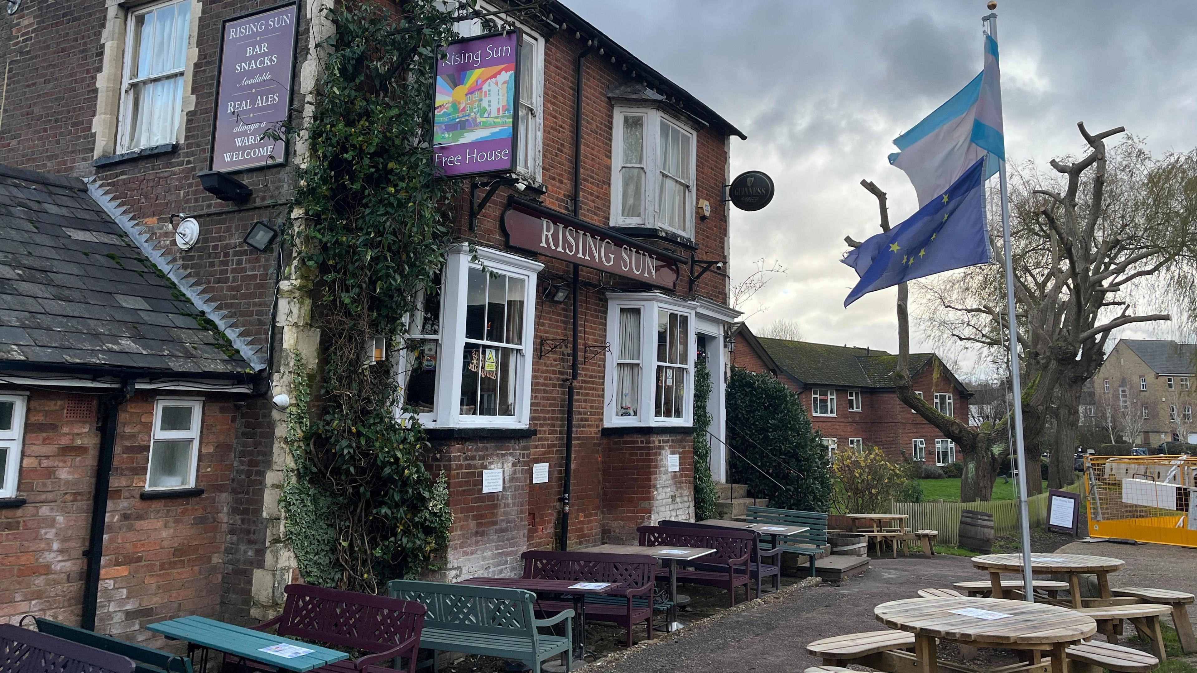 Outside of a pub with tables and benches and a European Union flag flying with houses in the background.