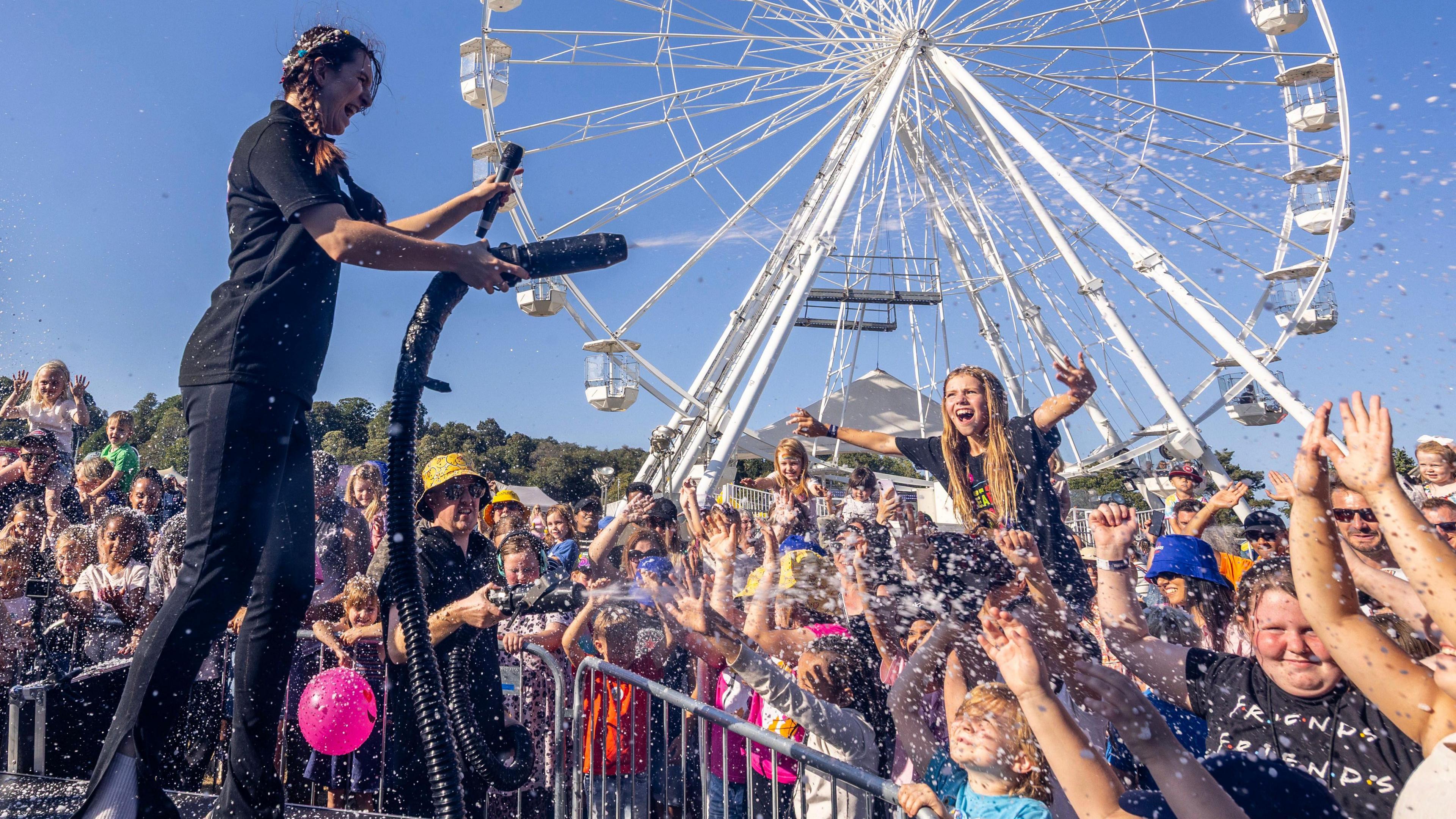 Many people having fun during an event with a big Ferris wheel in the background. A woman is pointing a foam machine at a young crowd as they smile.