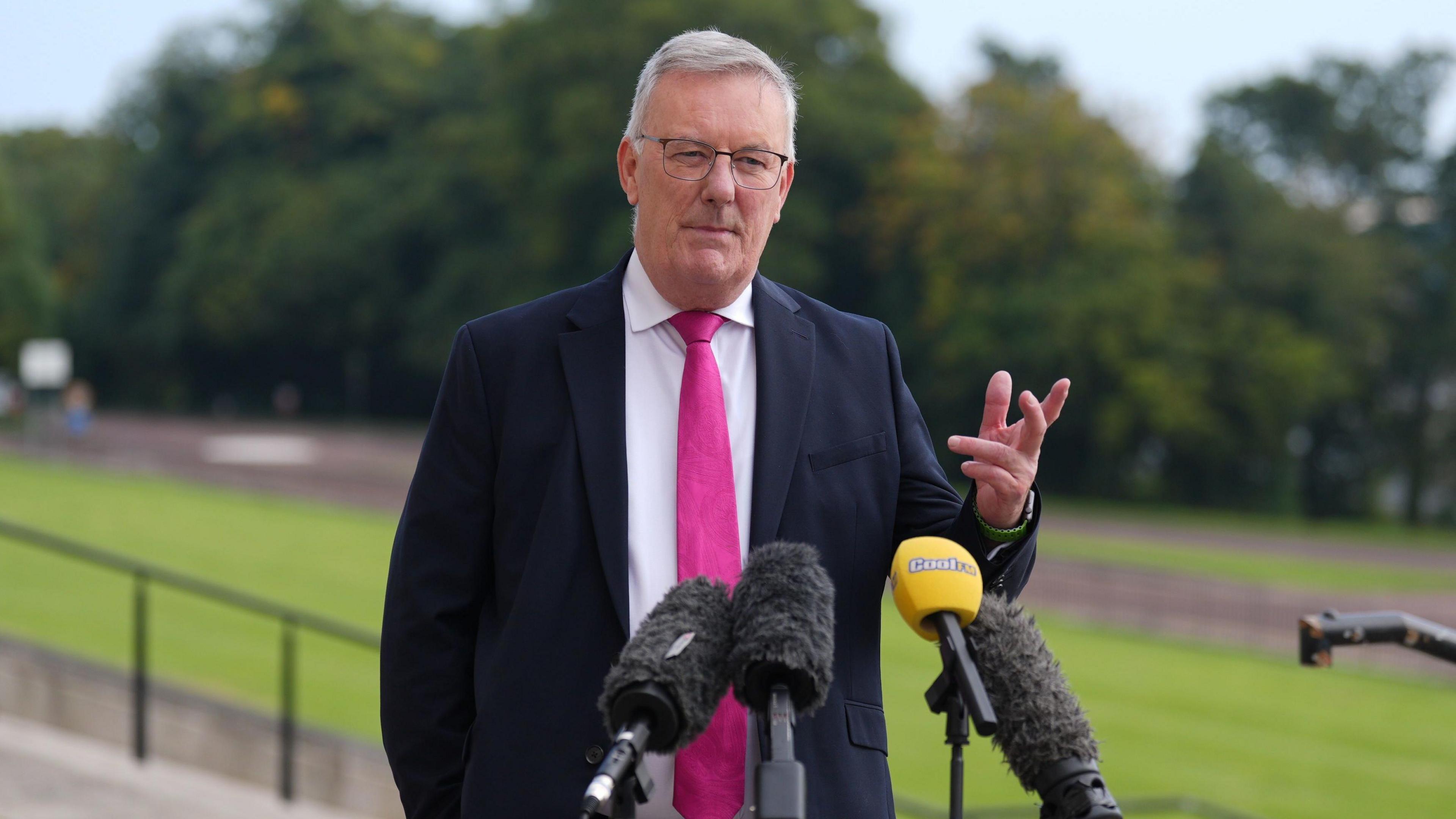 Mike Nesbitt standing speaking at microphones with one hand out in front of him and the other behind his back. He is wearing a suit and tie. Behind him trees and grass. He is speaking outside stormont - but the picture is not from today - it is an old picture. 