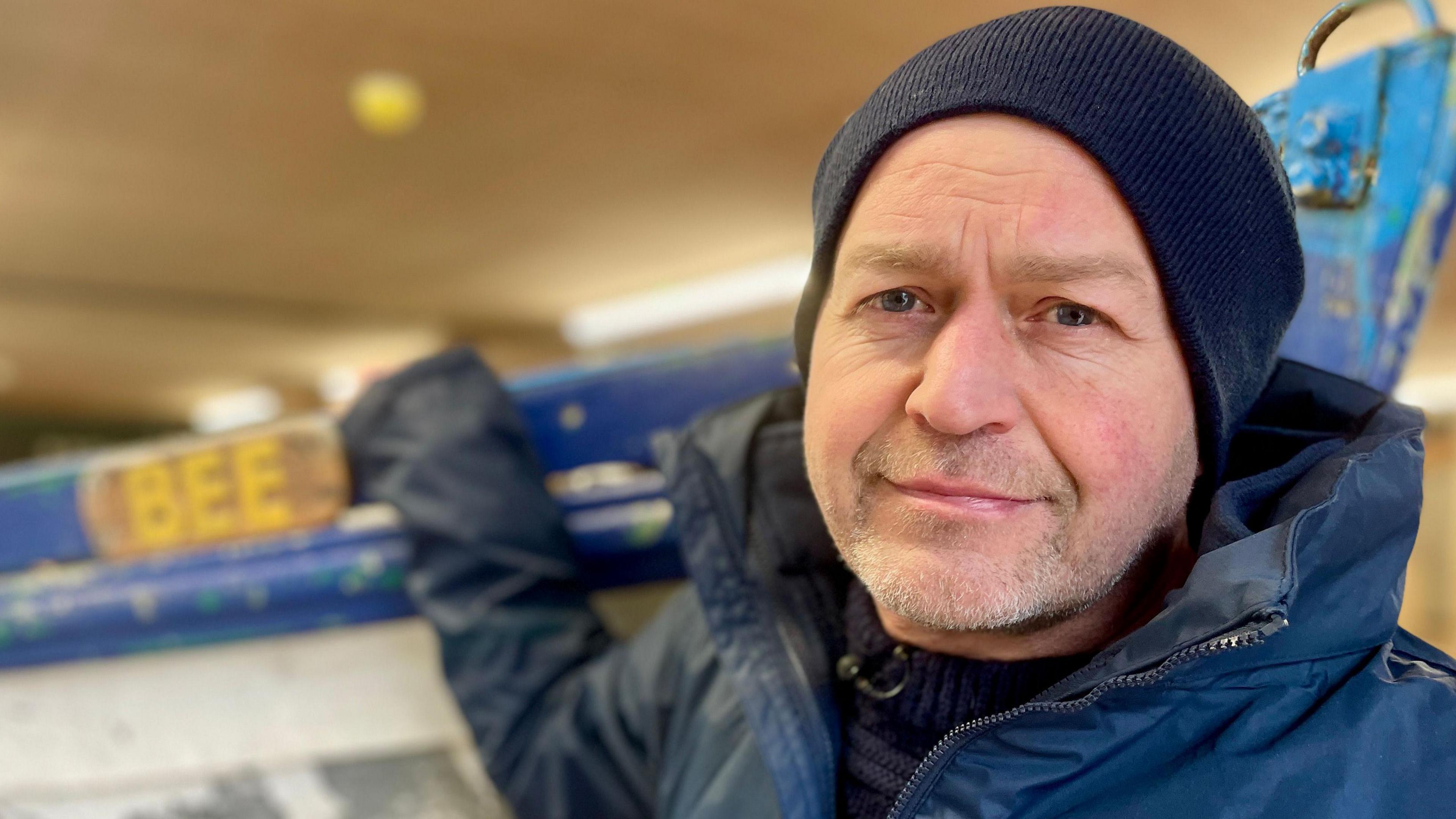 A man with a blue woollen hat and blue jacket faces the camera within a workshop with a sailing boat behind him