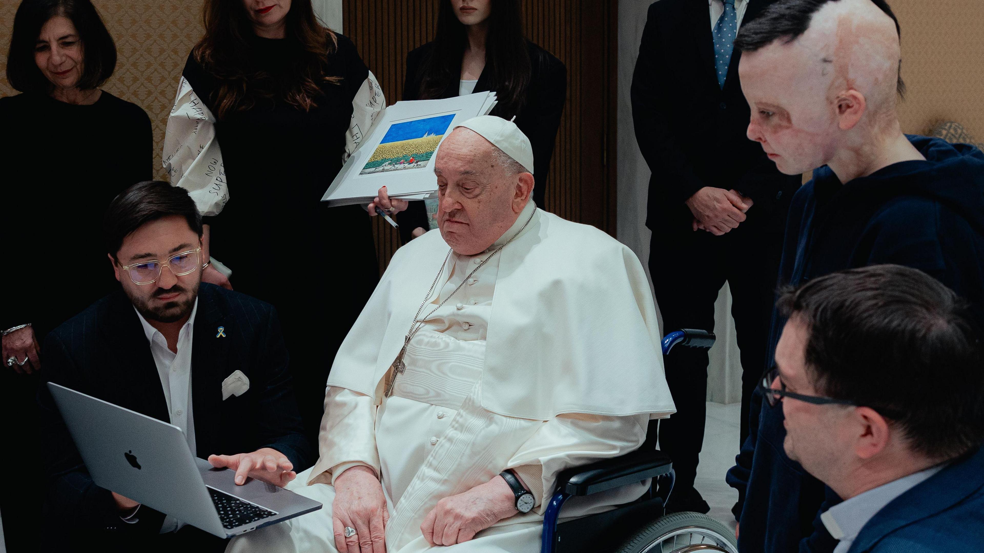 The Pope, surrounded by people, looks at a laptop being held by a man beside his wheelchair