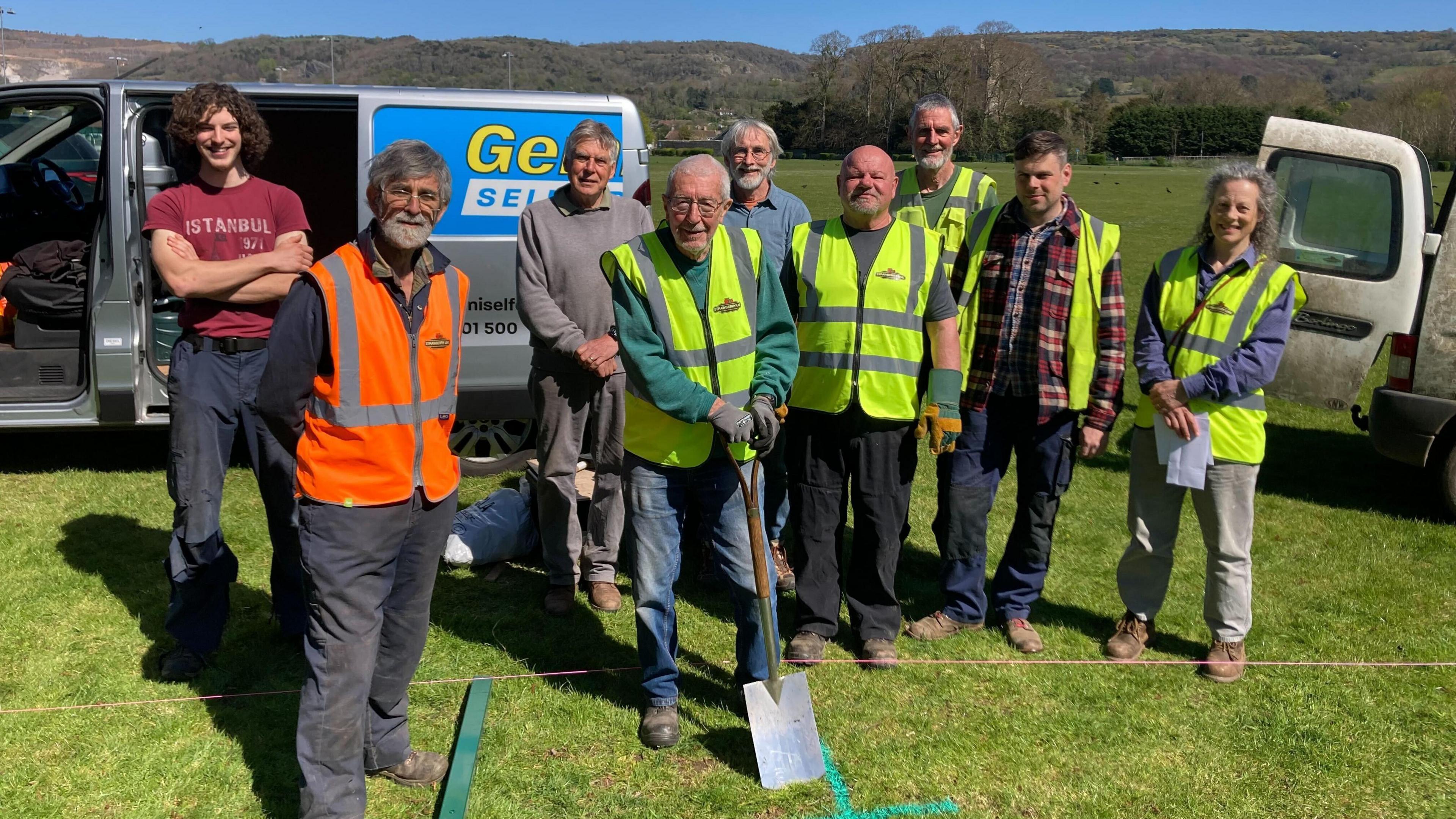 Strawberry Line volunteers beginning construction on the New Active Travel Route at Kings Academy in Cheddar. Some of them are wearing hi-vis jackets. They are all standing in a field and a man at the front is holding a spade.