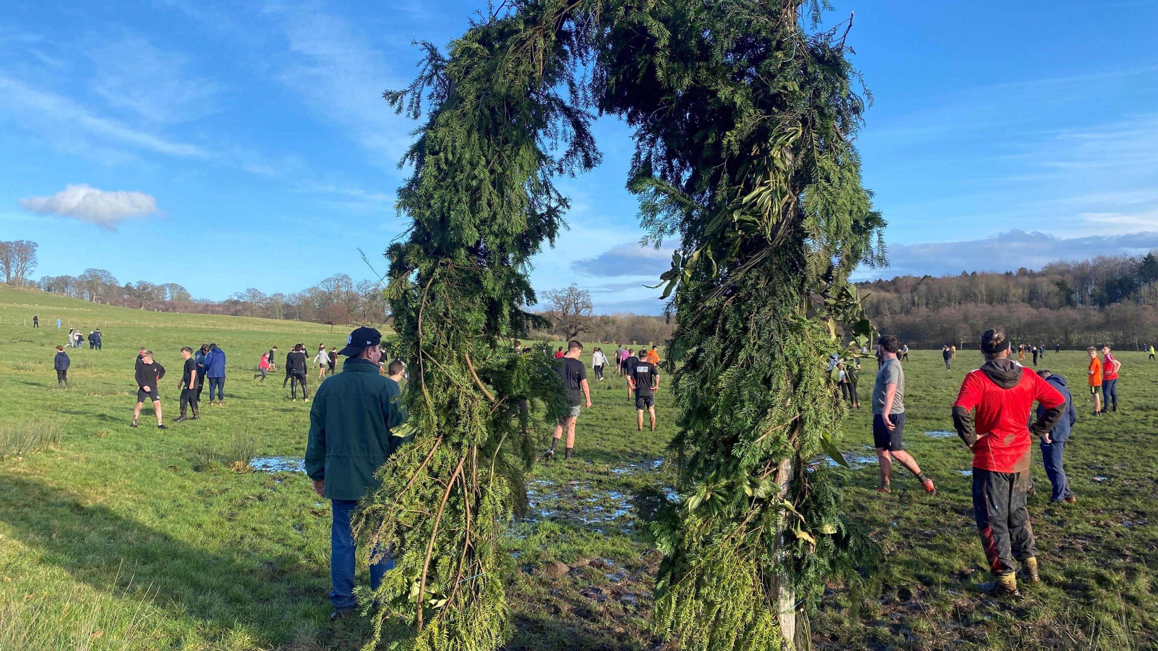 About 100 people are playing football on a large muddy field. In the foreground of the picture is a tall, horseshoe-shaped structure covered in ferns, known as a hale.