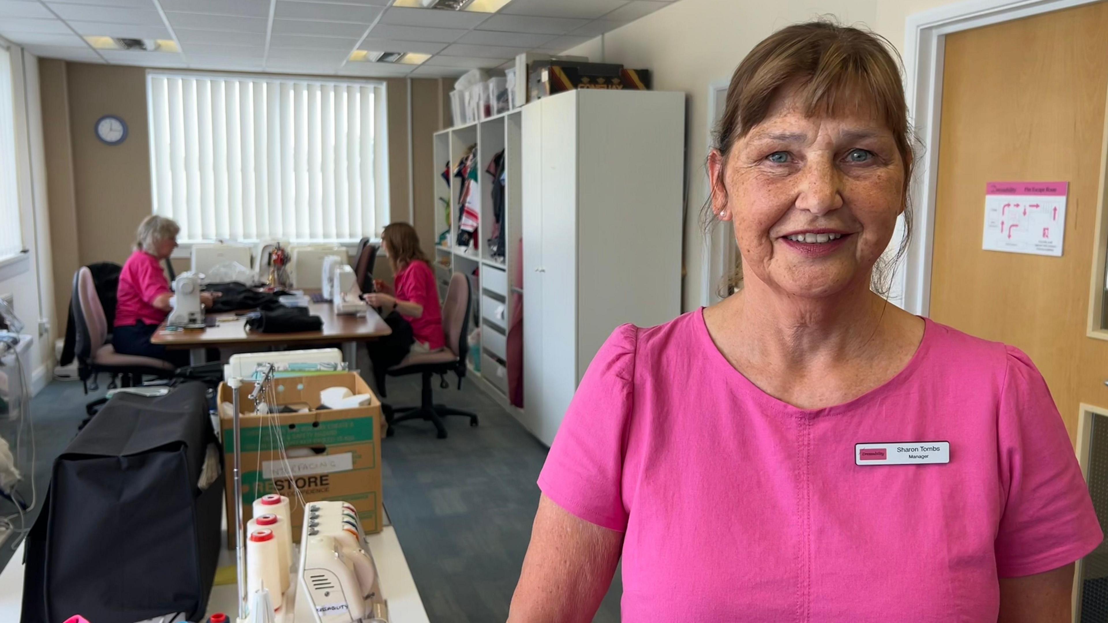 Sharon Tombs is wearing a pink top and a name badge and smiling at the camera. In the background two women are working on sewing machines.