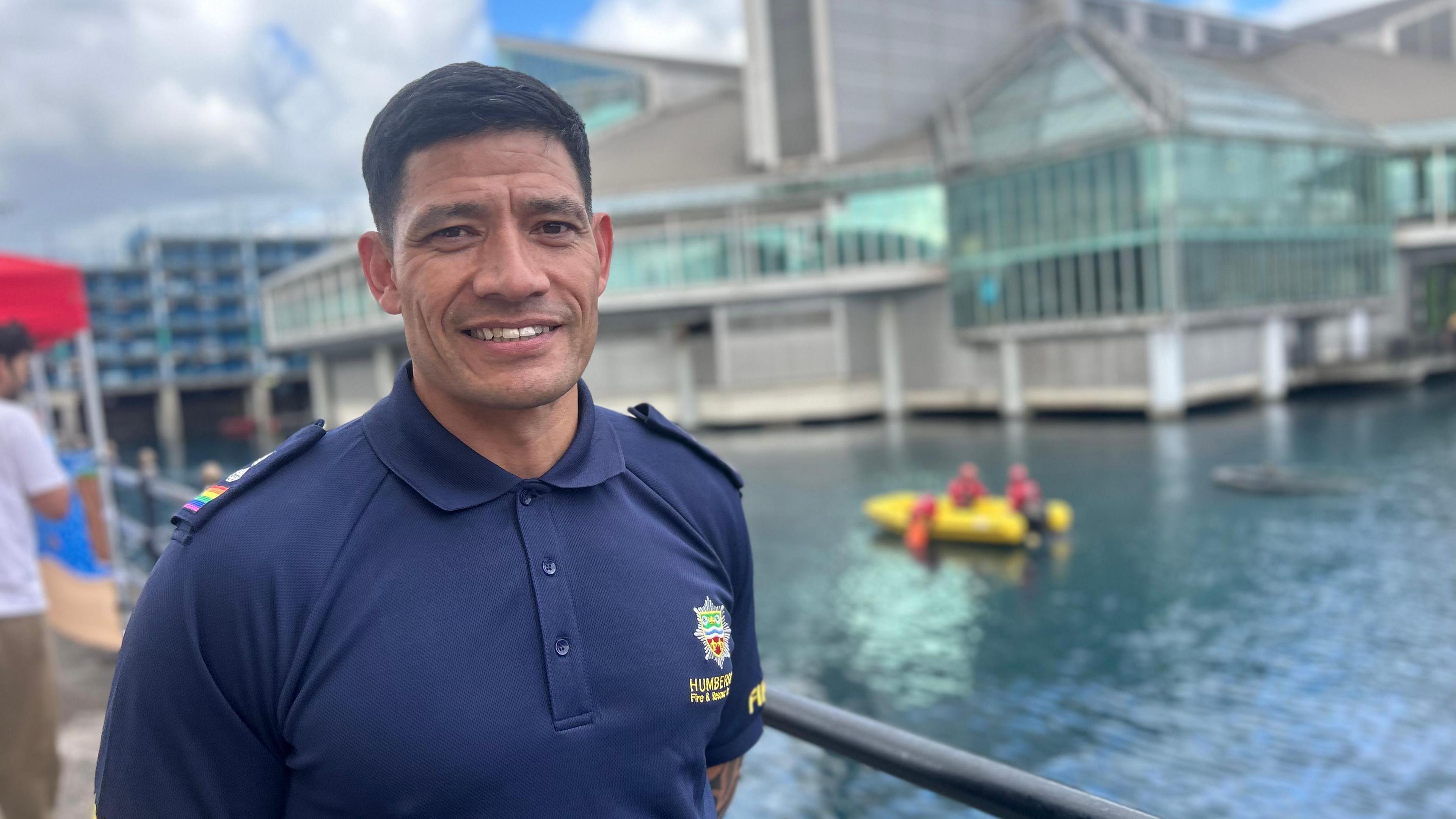 A man smiling in a blue fire and rescue polo shirt stood next to Princes Quay shopping centre with a yellow rescue boat training behind him