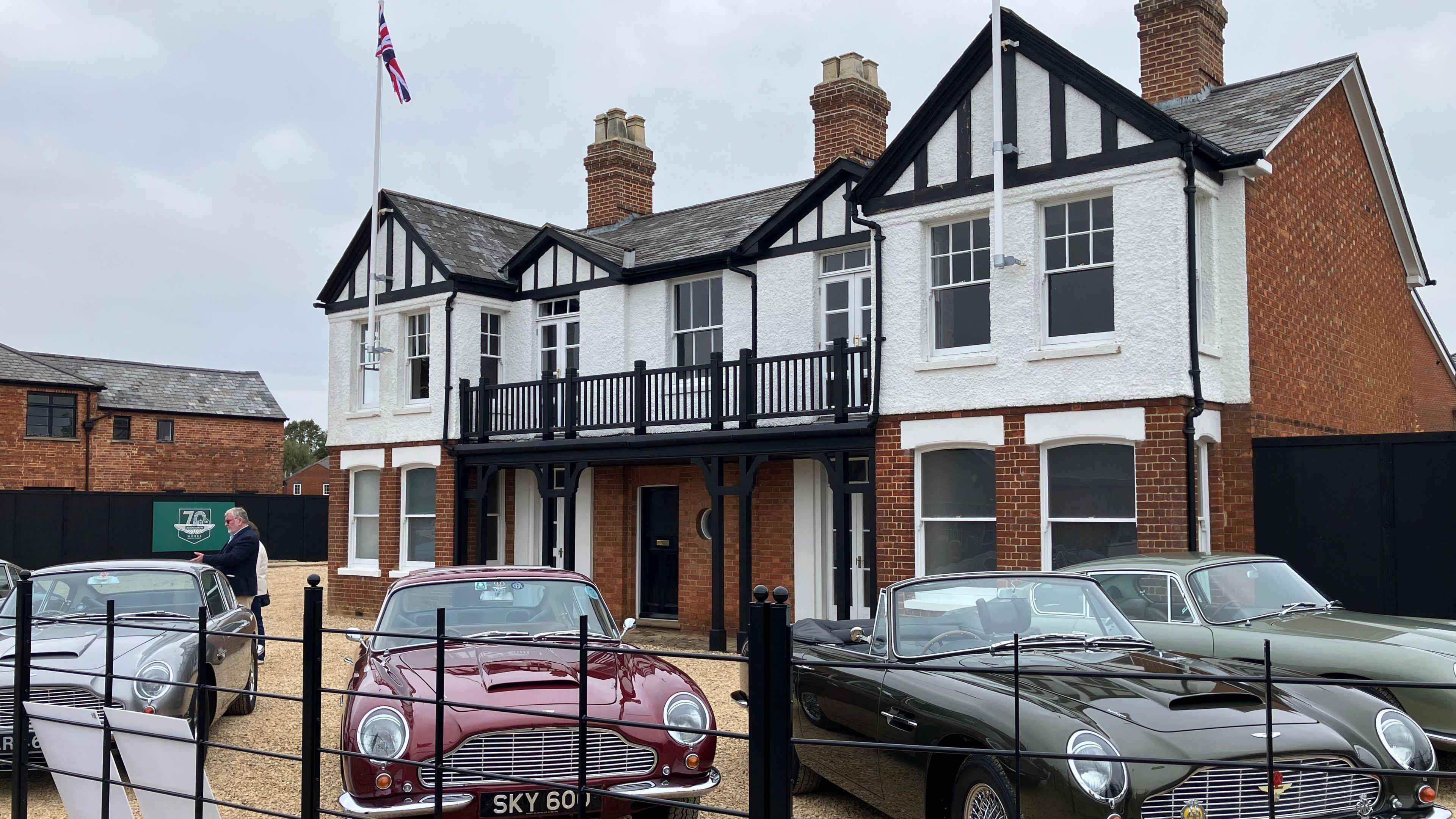 A building that looks like a large house can be seen. It is partly red brick and partly painted white with black facias. It has a grey slate roof. In front of the building on a large gravel driveway four Aston Martin sports cars can be seen parked up, pointing towards the camera. In front of them is a black metal fence that you can see through.