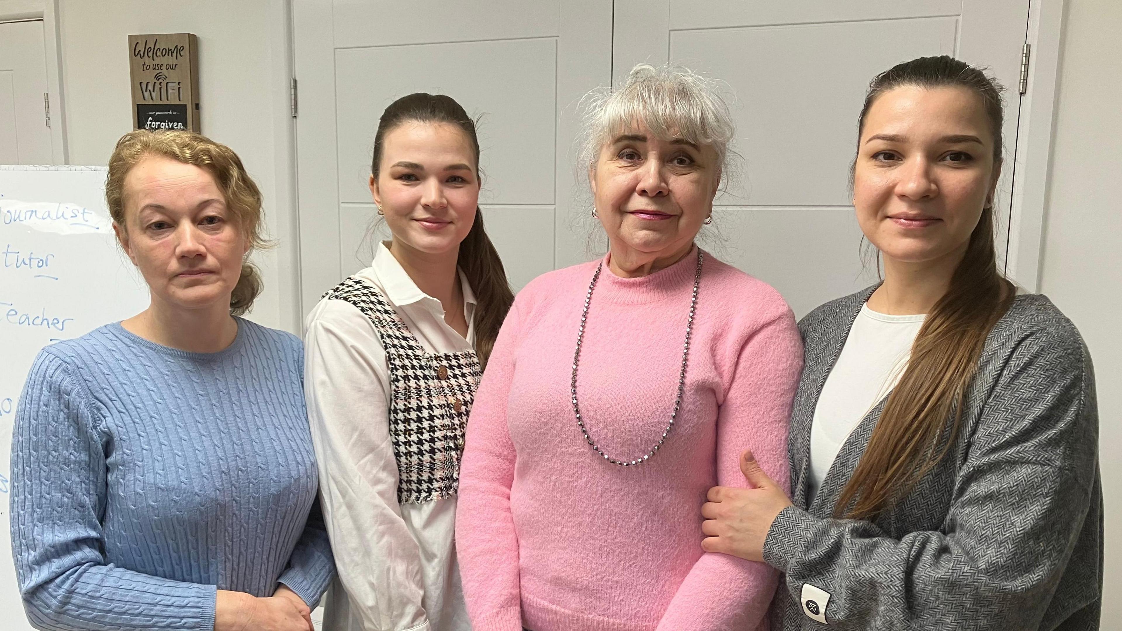 A picture of four ladies standing in front of a white board. On the left is a woman with blonde hair in a bob with a light blue jumper and jeans. Next is a woman with long light brown hair who has a white tunic and a chequred crop top. Next to her is a woman with grey hair who has a pink jumper on and blue jeans. And then on the right a woman with long dark brown hair wearing a gray cardigan and black trousers.