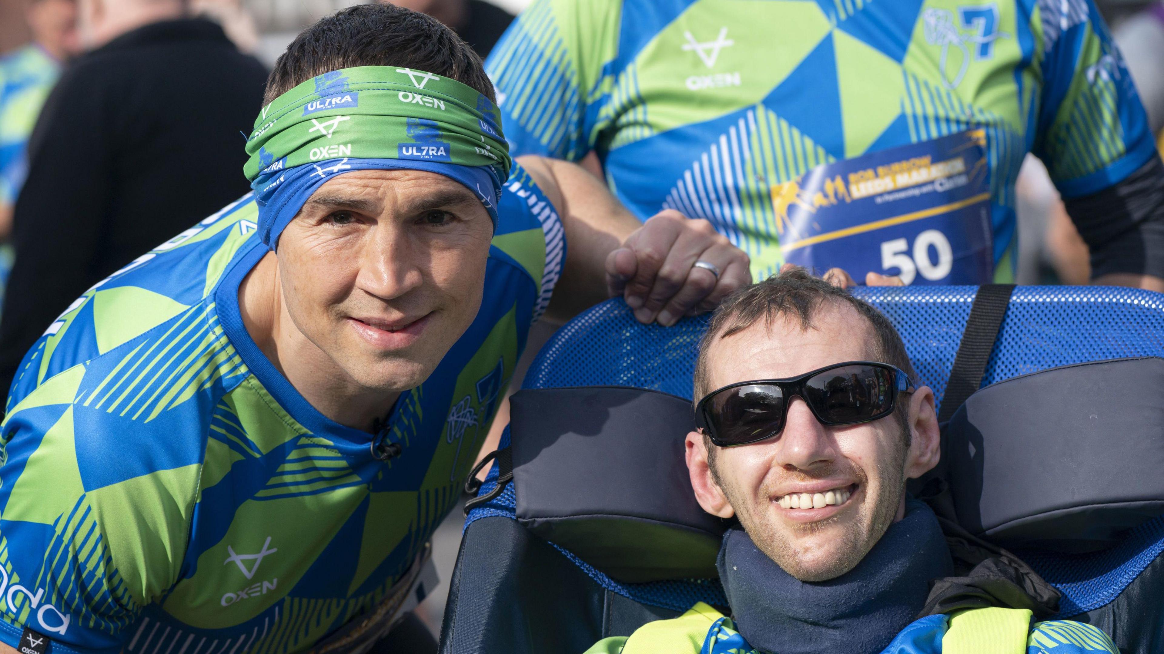 Kevin Sinfield stands with Rob Burrow. He wears a blue and green sports top and bandana. Rob is seated on a motability scooter and wears sun glasses and smiles