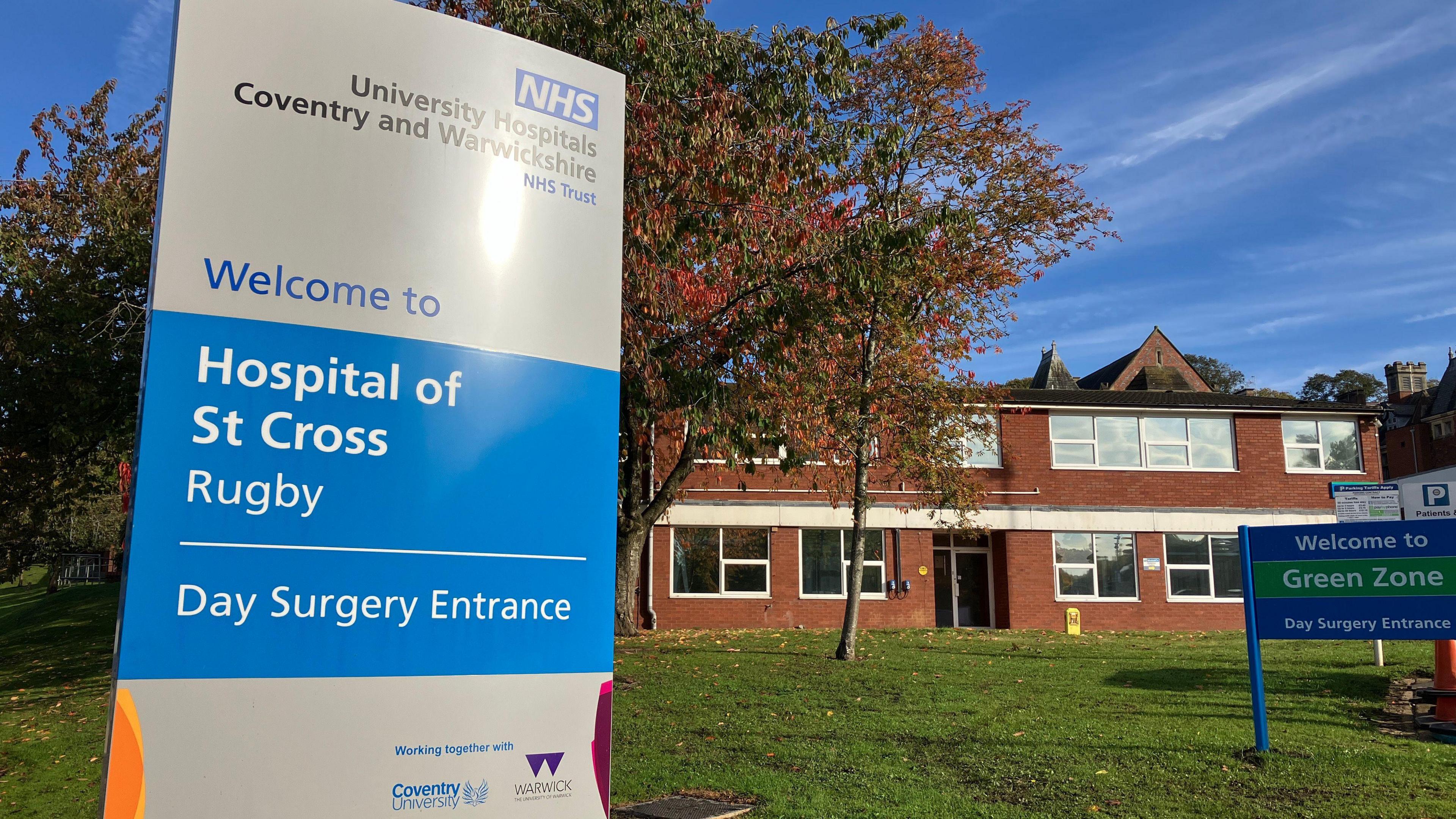 A blue and silver NHS sign that says Welcome to Hospital of St Cross Rugby, in front of a red brick building and a tree with autumnal leaves