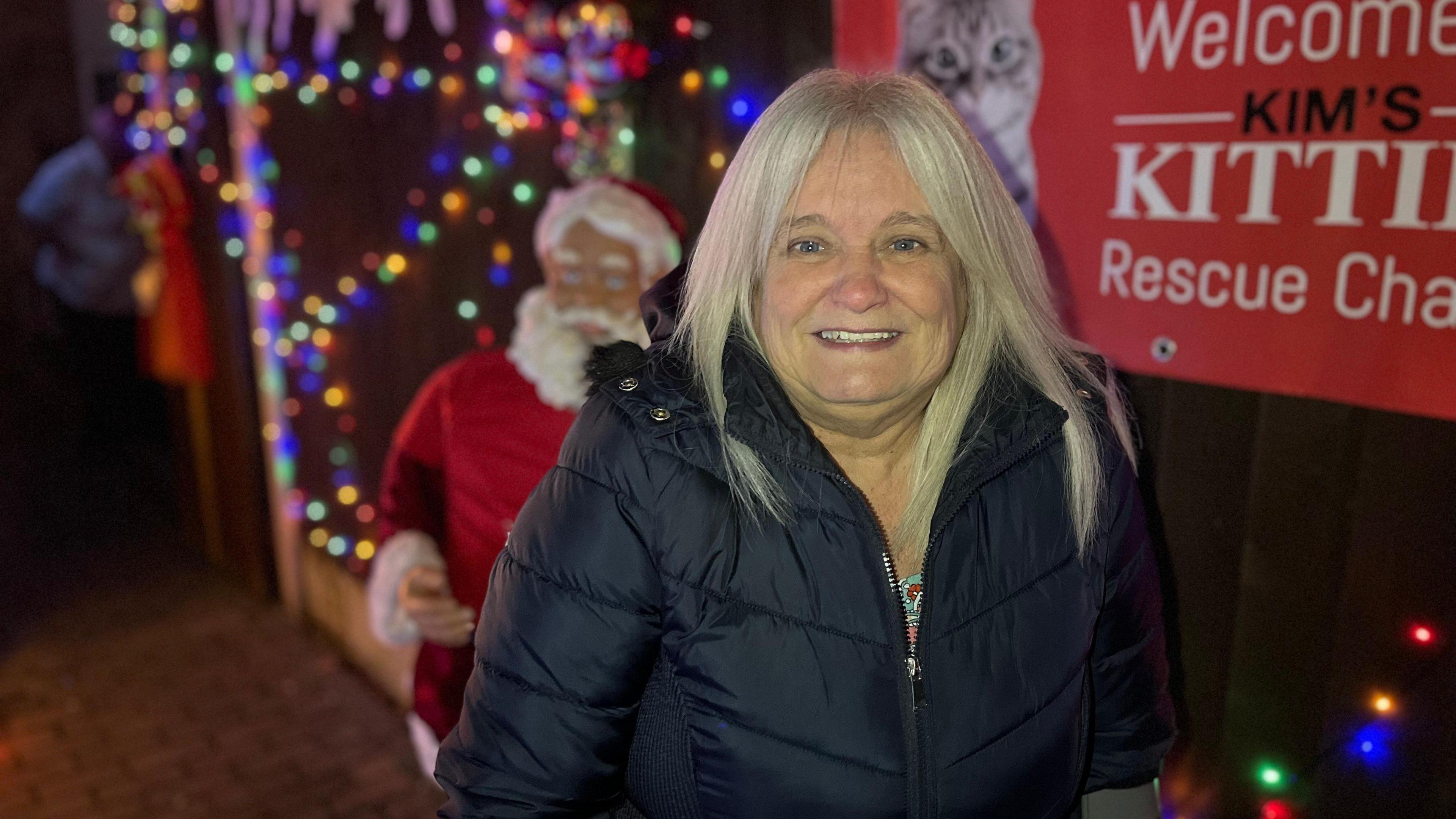 Debbie Saunders with long silvery-blonde hair smiles at the camera while wearing a warm winter coat. In the background is a poster for a charity and a Father Christmas figure.