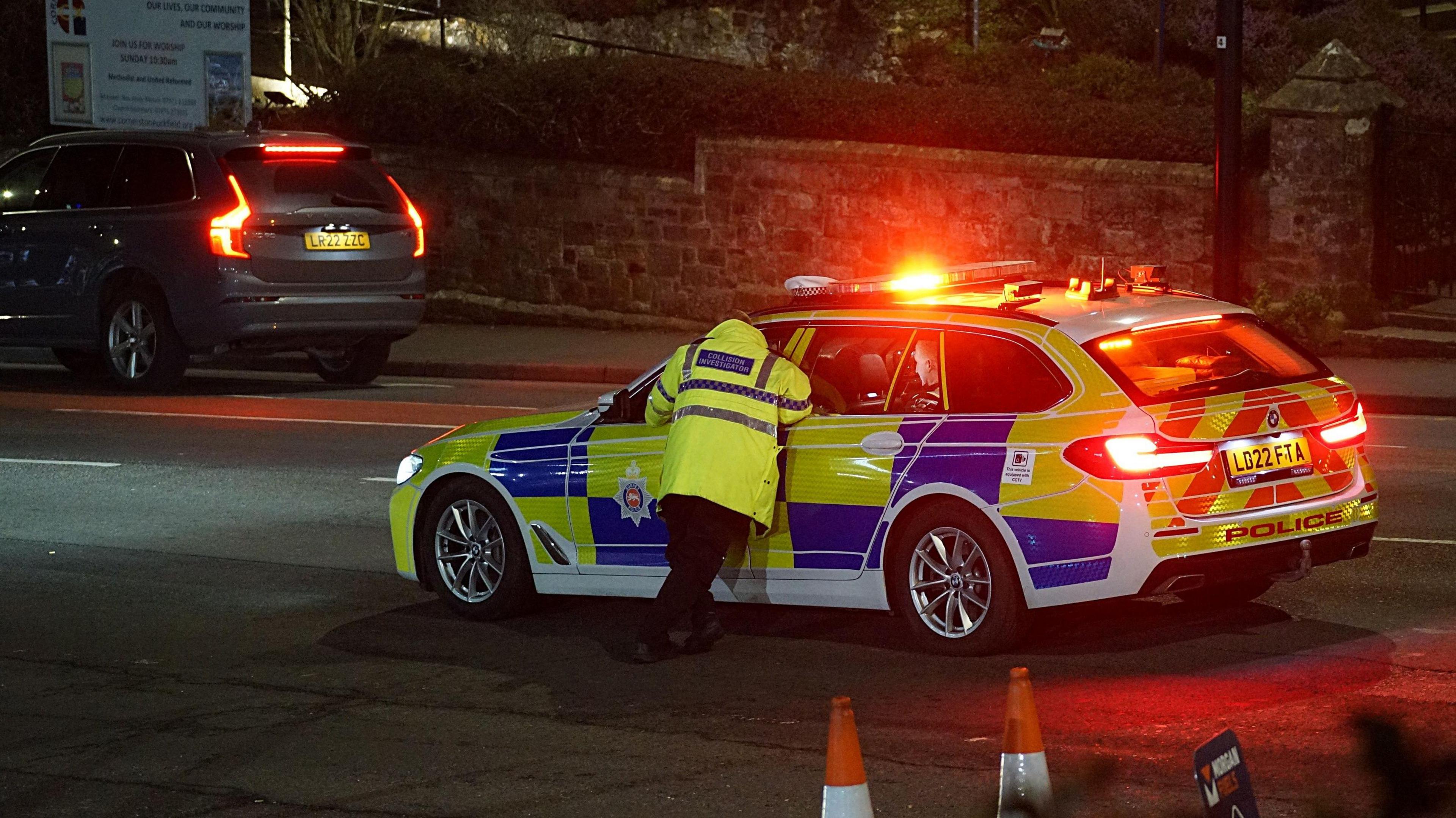 A police officer leans into the window of a police vehicle, which has flashing lights