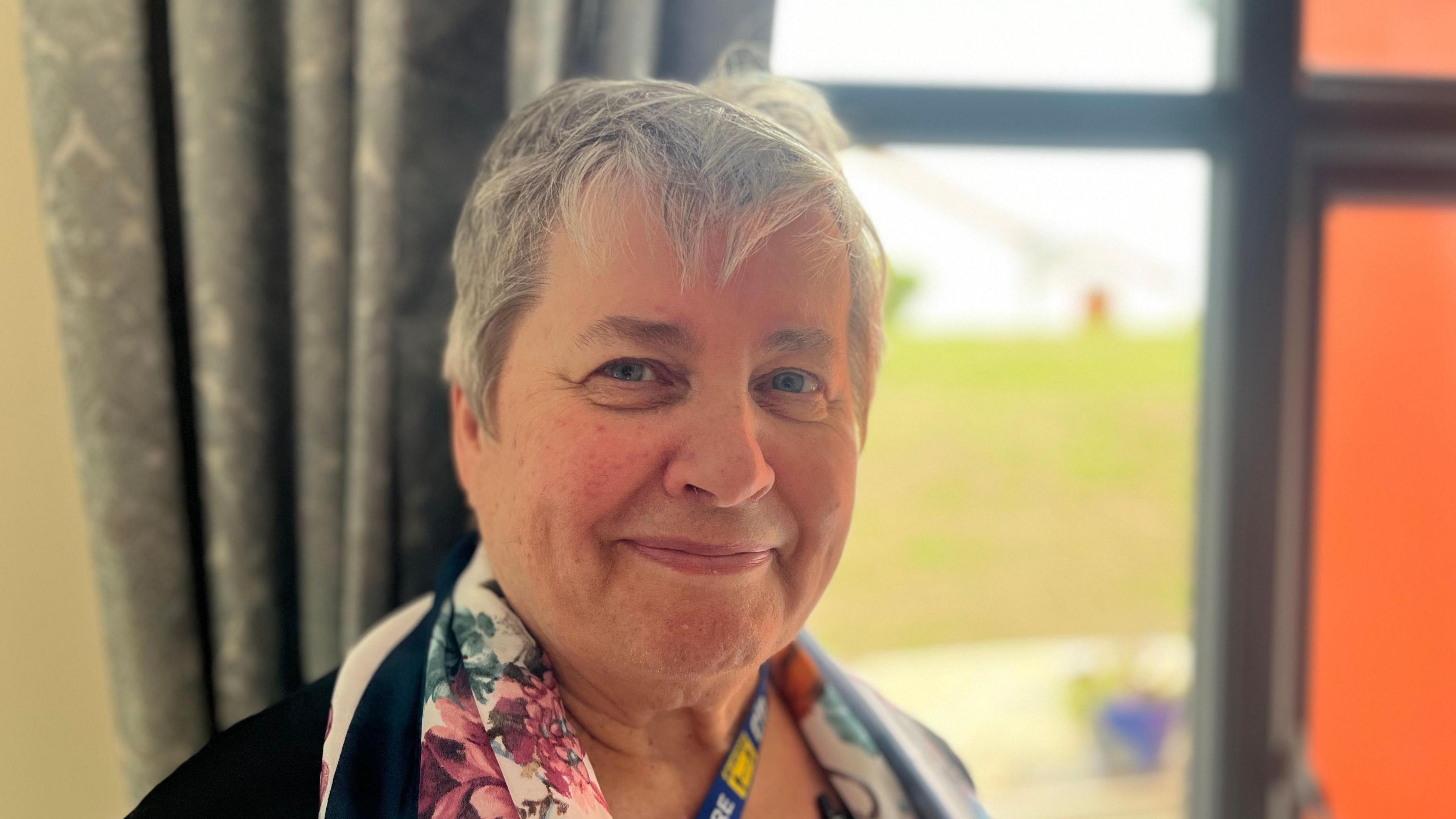 a smiling woman with short grey hair looking into the camera with a window behind her