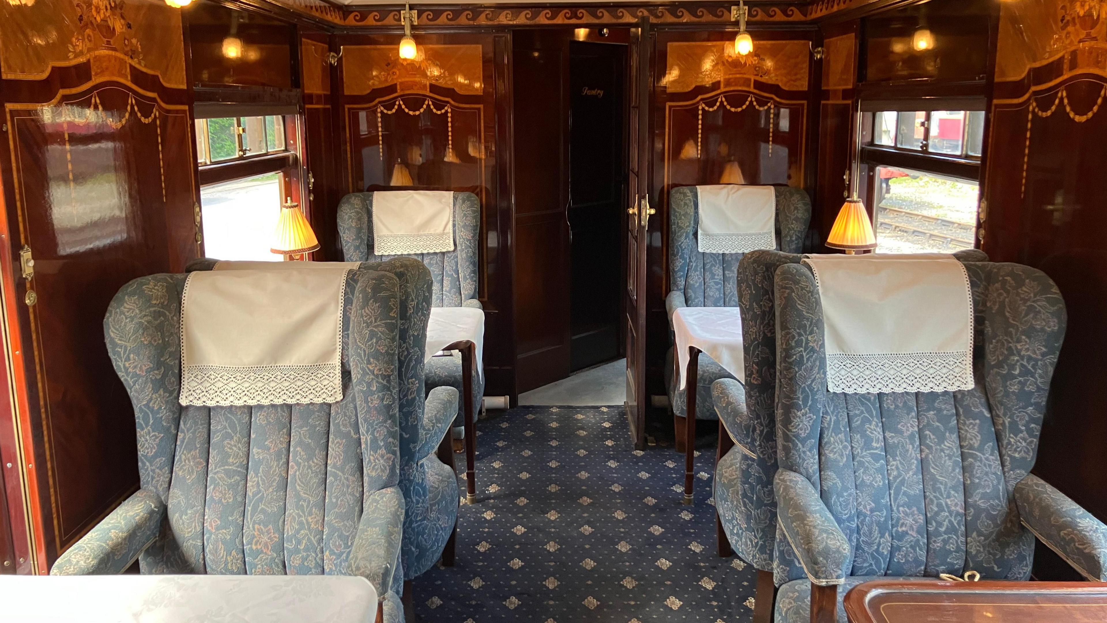The inside of the carriage. The seats are a light blue with a white flower pattern. The carpet is a navy blue with white diamonds. The walls of the carriage look like they are made of wood. There are tables in front of the chairs and and there are lamps on the tables.