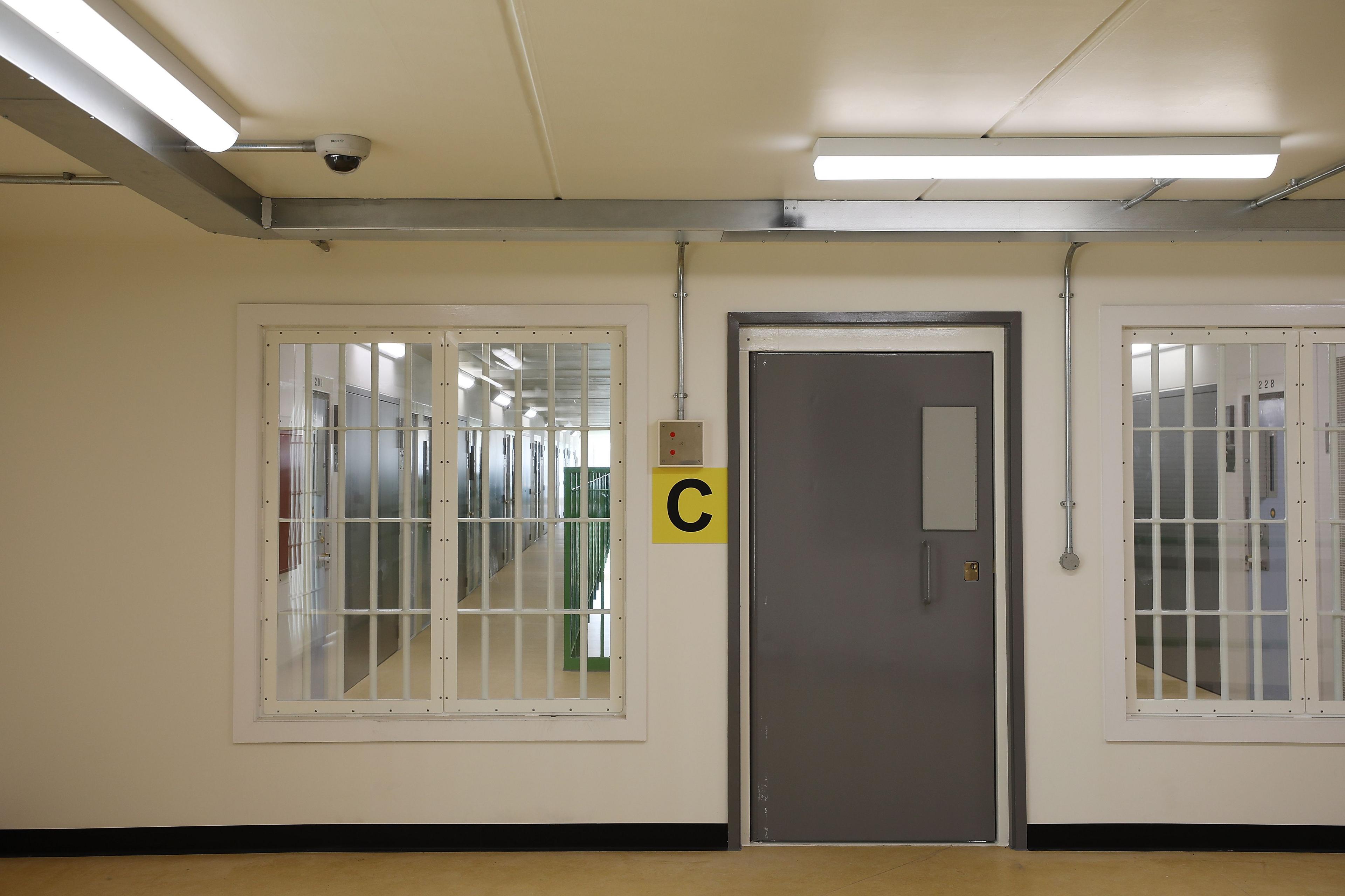 A cell block can be seen with the letter 'C' hanging by the door. Through windows either side of the door a corridor with a row of more doors can be seen.
