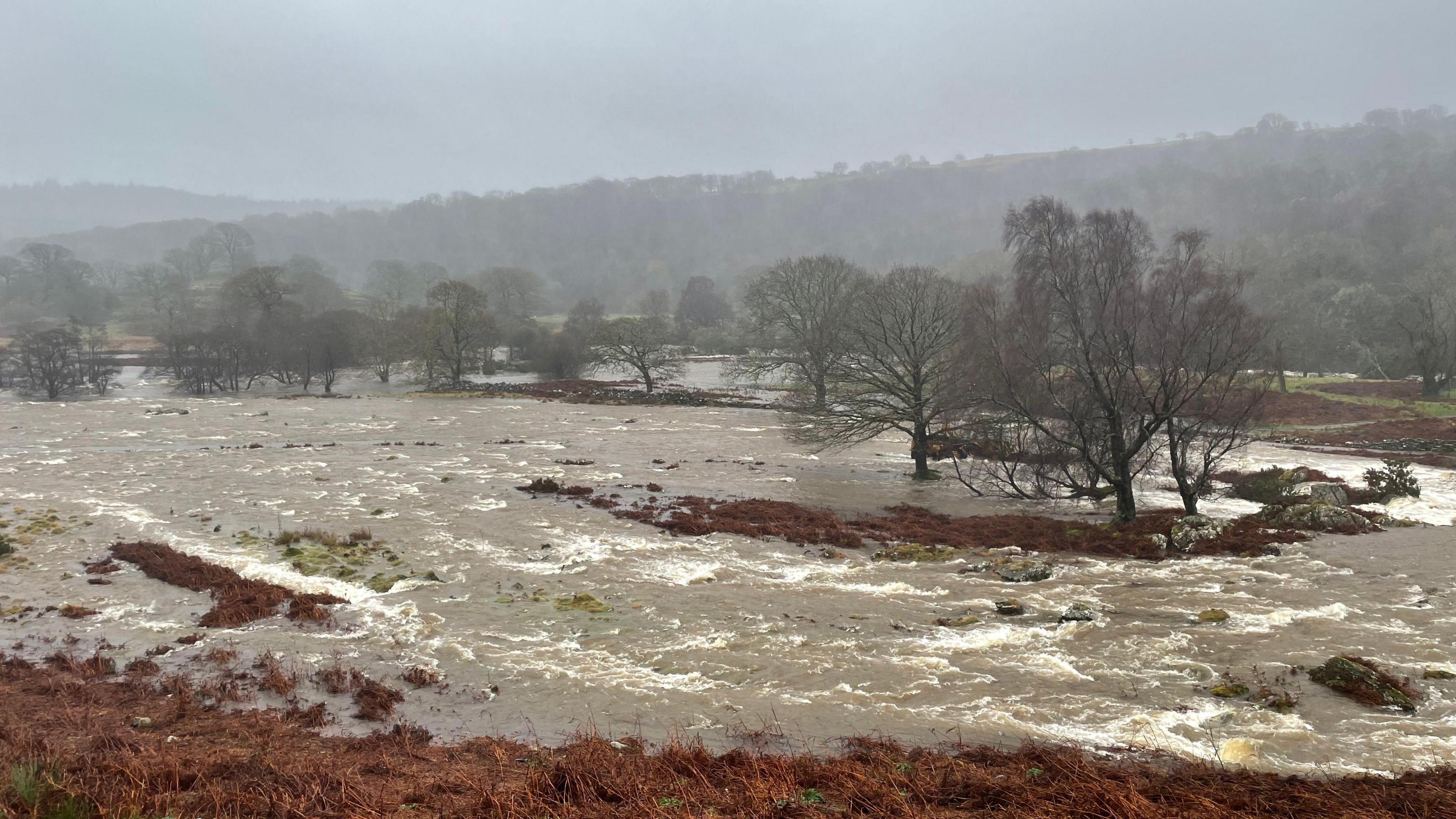 An area of the countryside in Wales with a full flowing river and trees caught in the wind