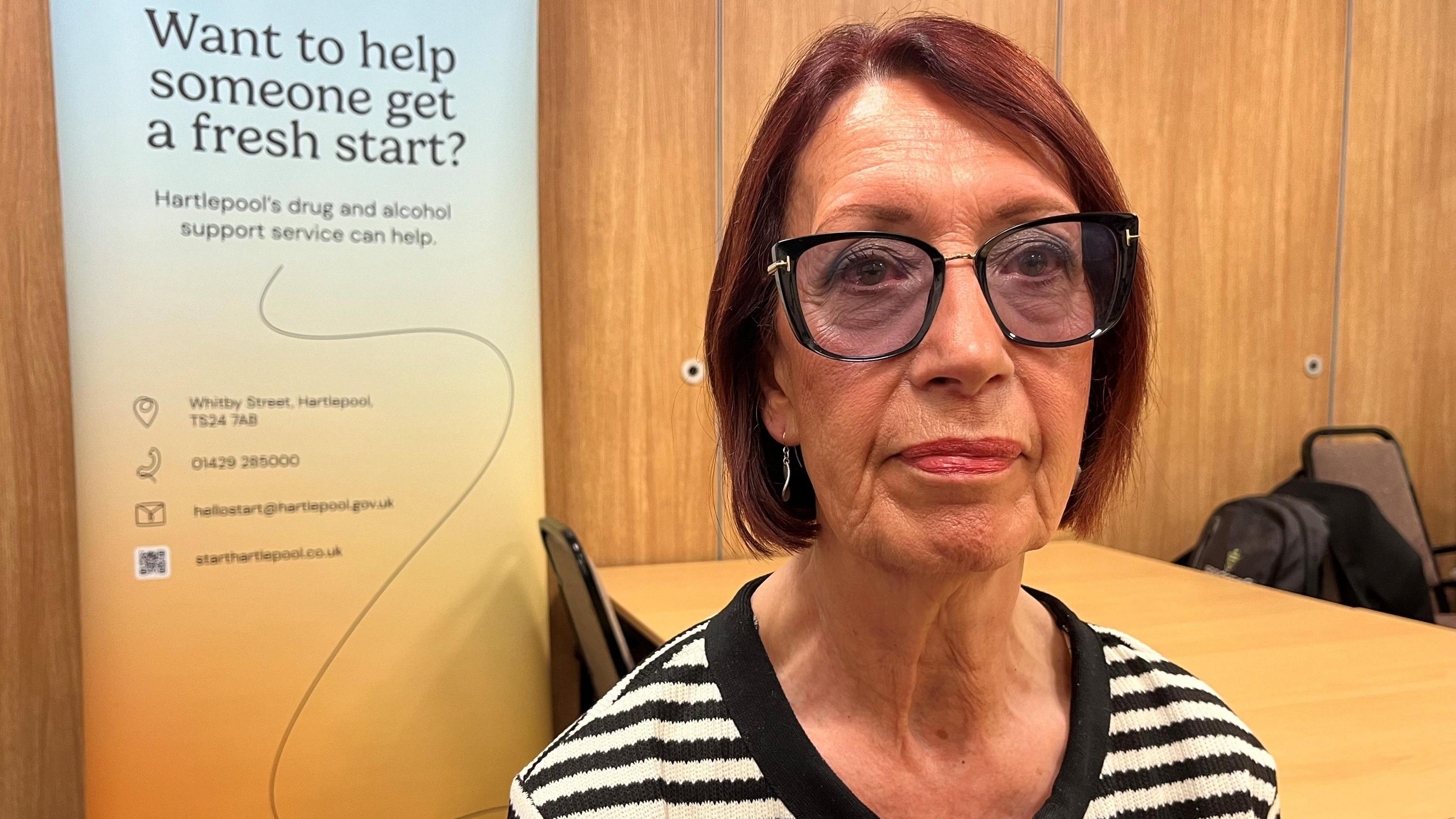 Brenda Harrison, Chair of Hartlepool’s Health and Wellbeing Board and Leader of Hartlepool Borough Council, sitting by a table, wearing glasses and a stripy top, with an advice banner behind her. 