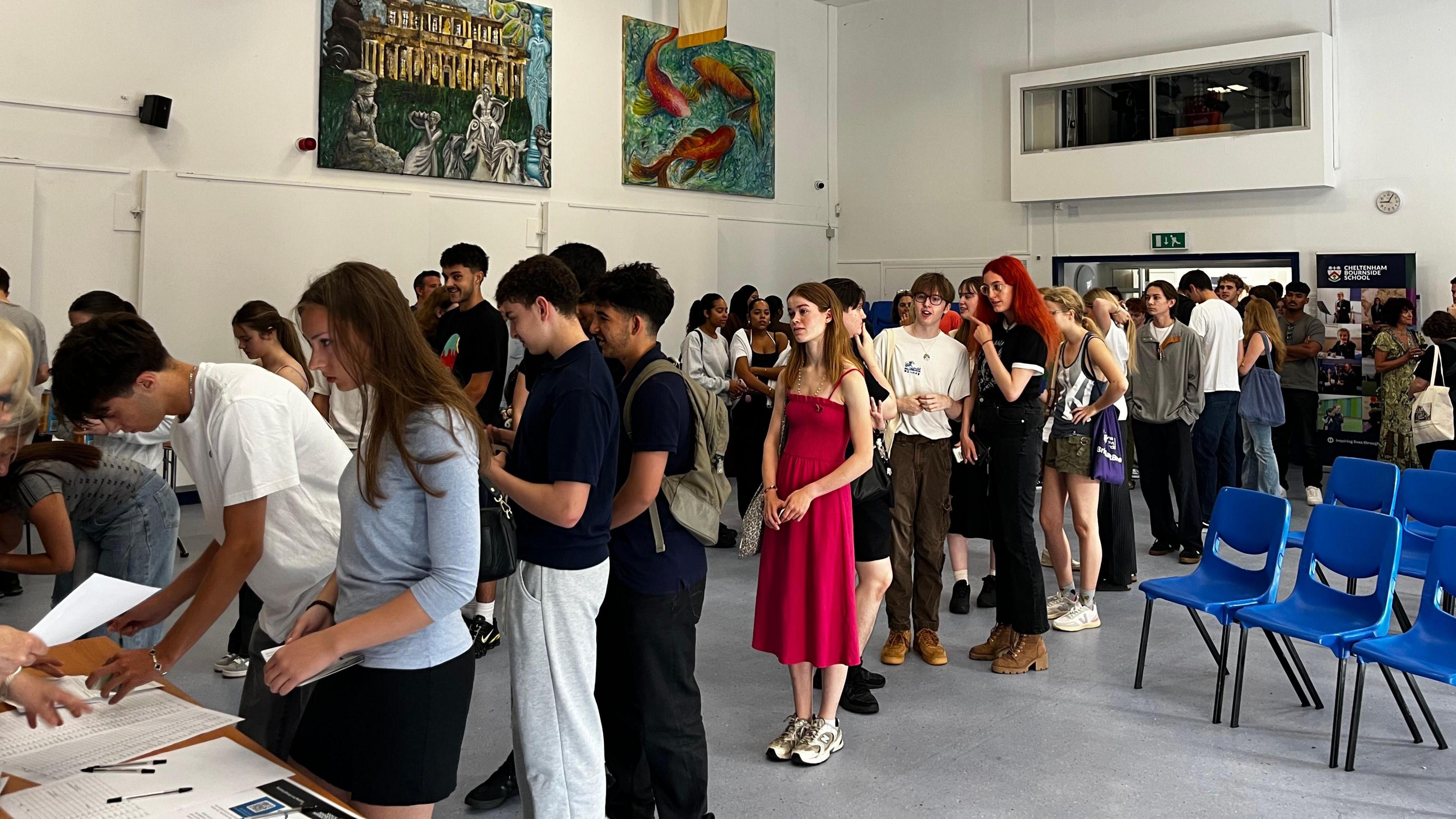 Students at Cheltenham Bournside School queuing up in a large white hall. They are waiting to get their A-Level results envelope from a white table at the front of the line. There are blue plastic chairs on the right and large canvas artwork on the walls above.