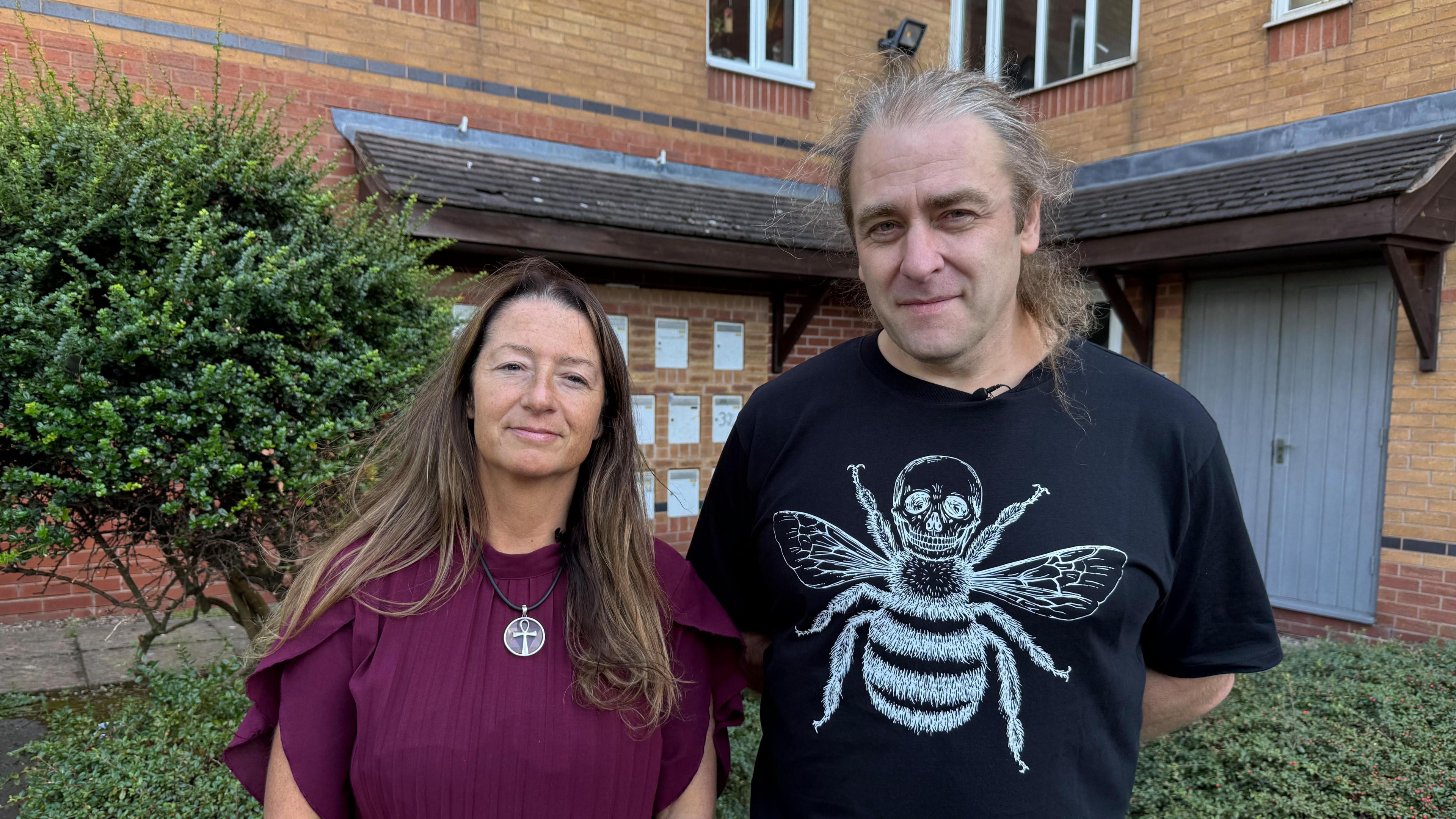 Neighbours Naomi and Rob standing next to each other and smiling. We can see there is an apartment complex behind them. Naomi is wearing a purple dress and a necklace with a cross on. Rob is wearing a white t-shirt with a skull and a bee on it.