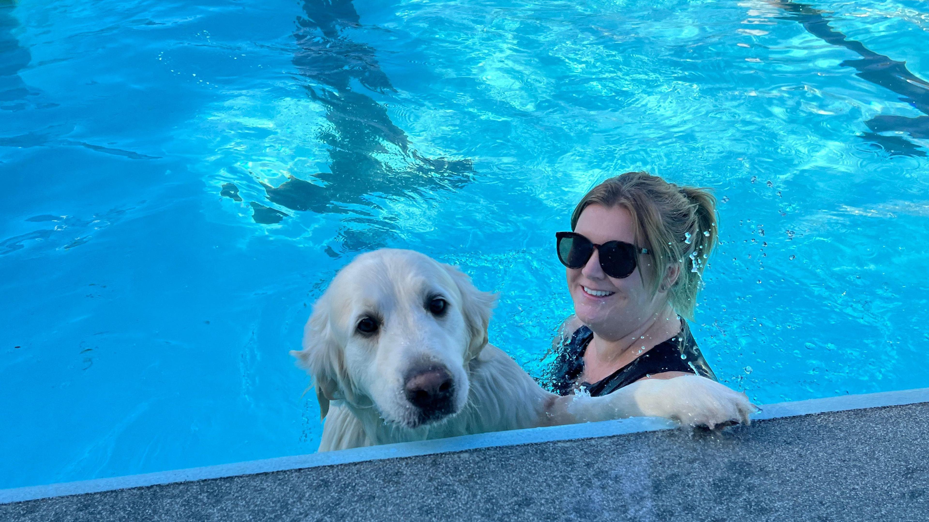 Katrina wearing a black sleeveless swimming costume inside the pool holding her white Golden Retriever George.