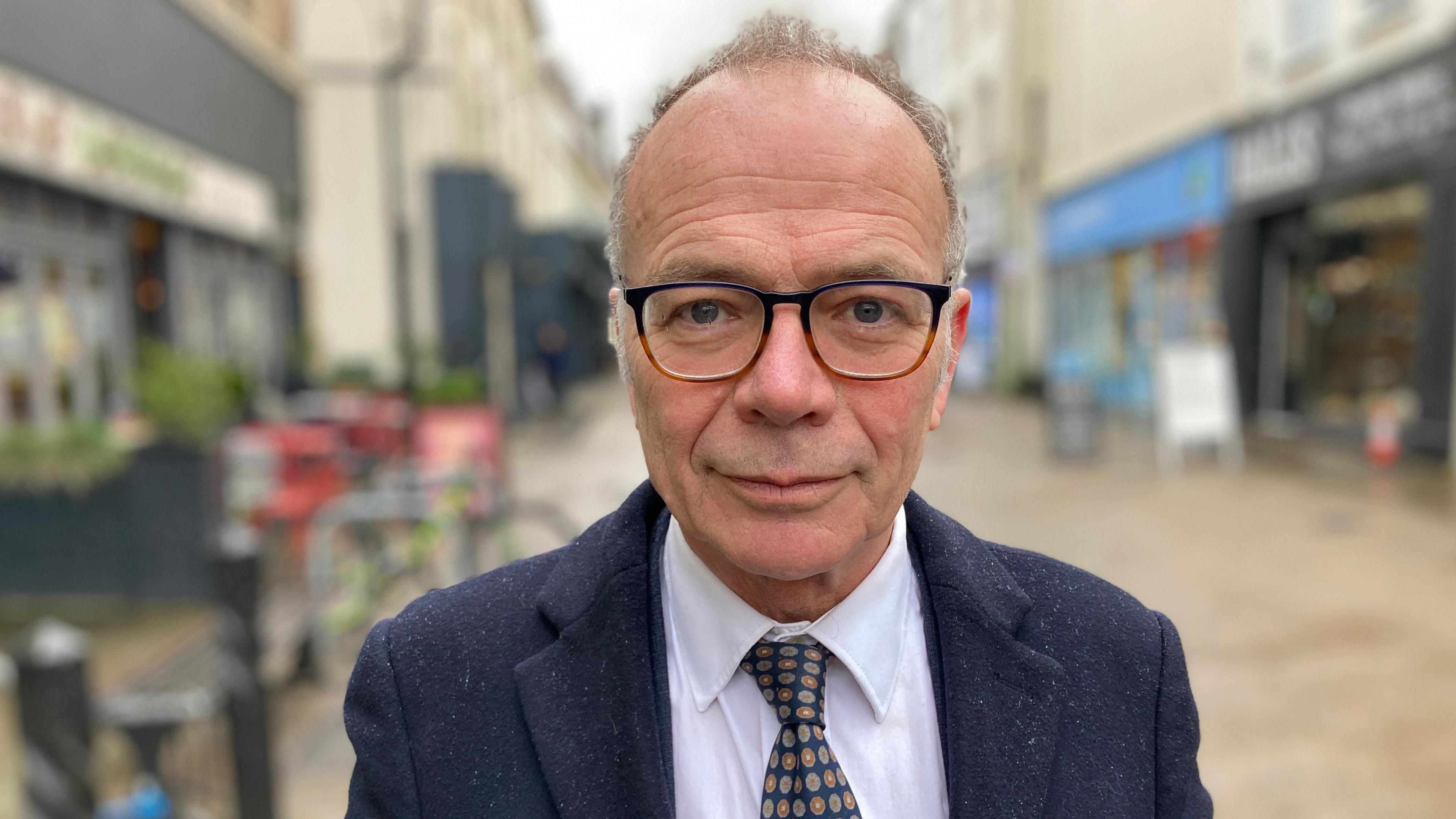 Dr Simon Opher - an older man wearing glasses, a suit, white shirt and spotty tie, stands on a shopping street in Stroud.