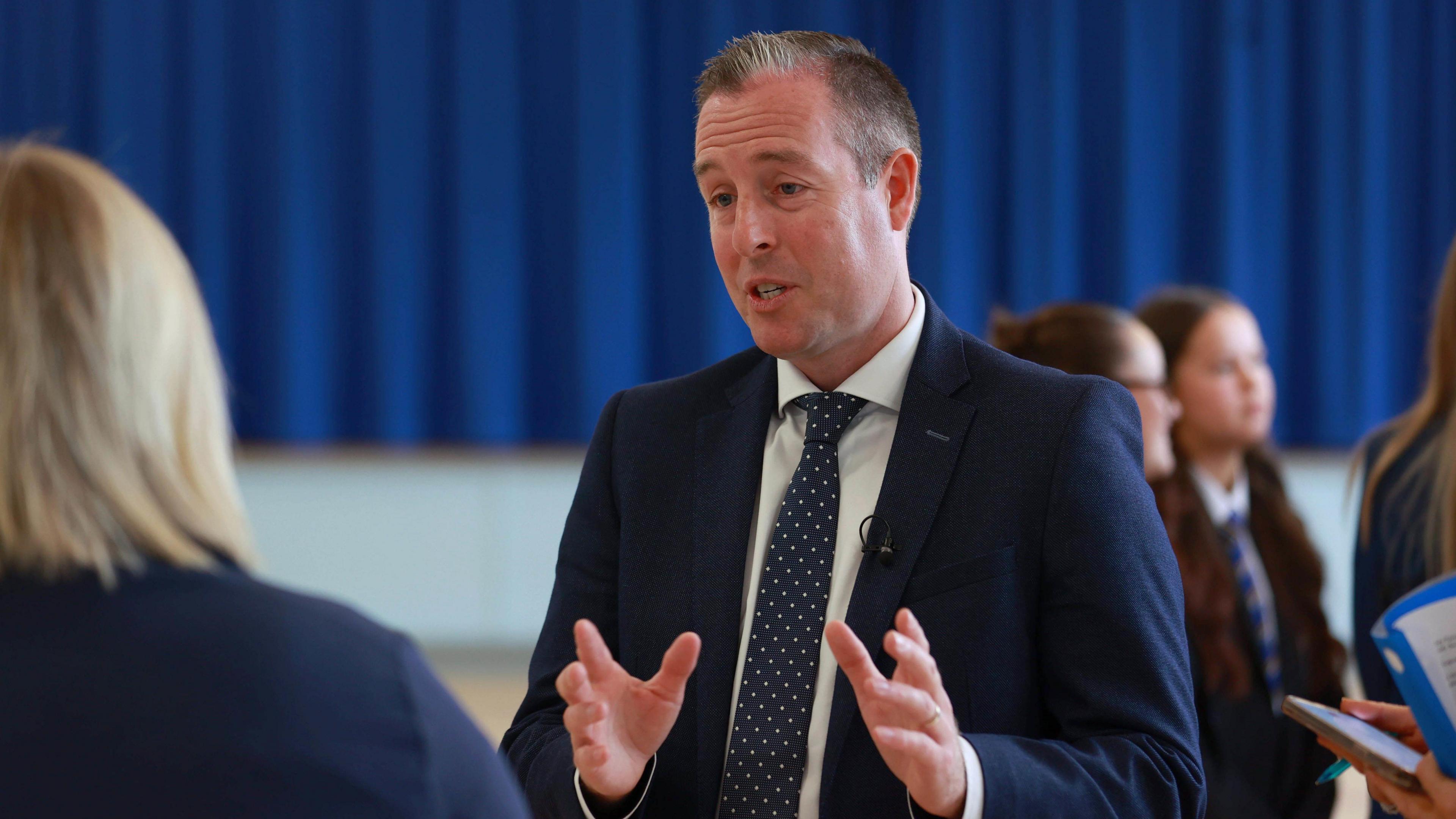 Paul Givan standing and speaking with his hands out in front of him. He is wearing a blue suit and tie. The person he is speaking to is facing away from the camera and is out of focus. In the background some girls wearing school uniforms. Behind them a blue curtain.