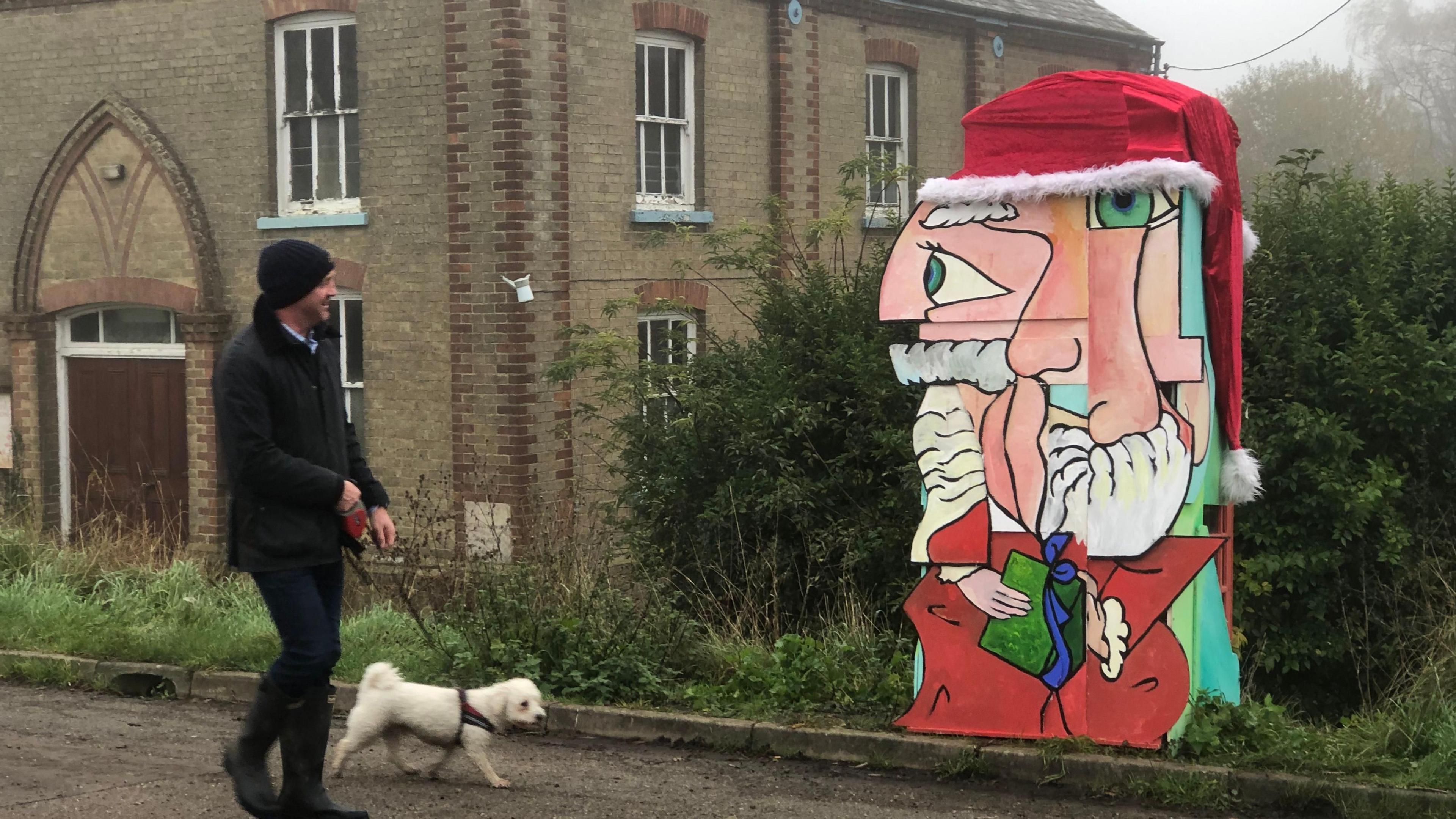 A model of a Santa face painted in the style of the artist Picasso. The eyes, nose and mouth are all in ususual places and it is wearing a red hat. A man is walking past with a dog, admiring the art
