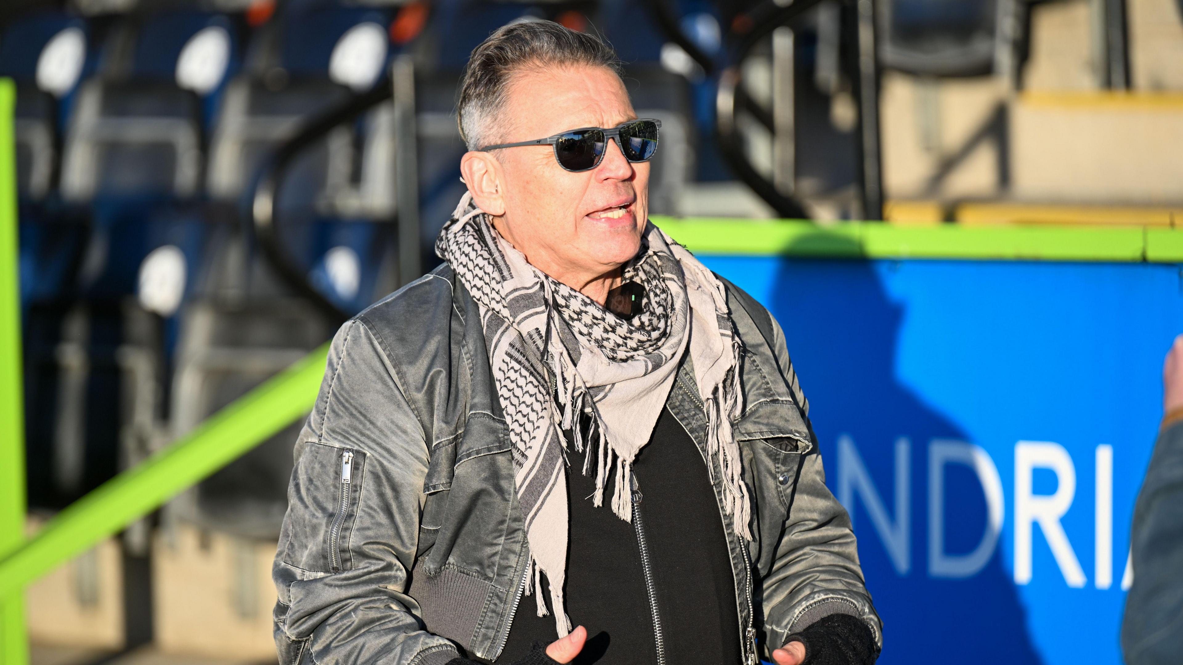 Dale Vince walking at Forest Green Rovers' Bolt New Lawn Stadium. He is wearing a grey coat, white, black and grey patterned scarf and black sunglasses