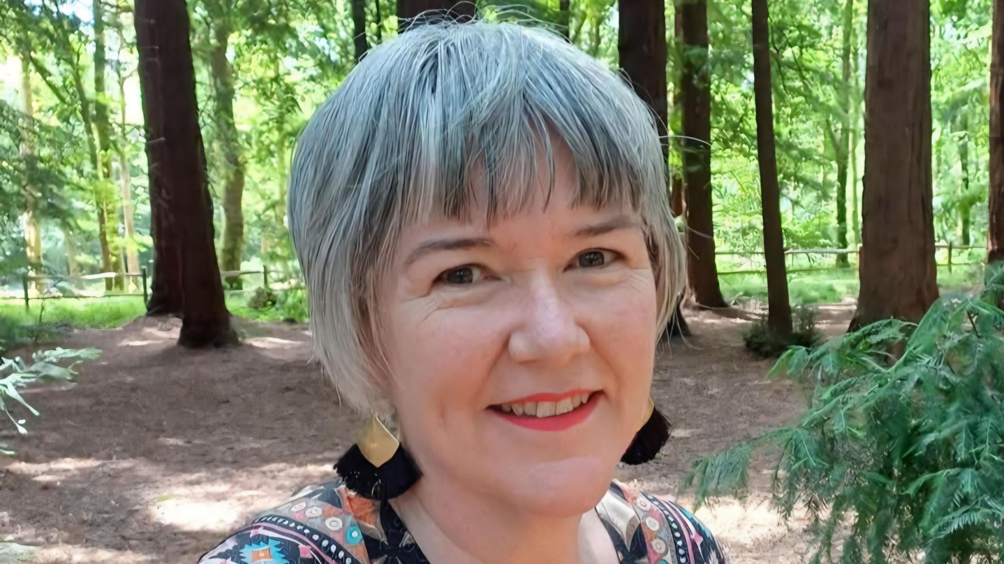 A woman with short grey hair, black and gold earrings, and a multi-coloured top, smiles as she takes a selfie in the middle of a wooded area.