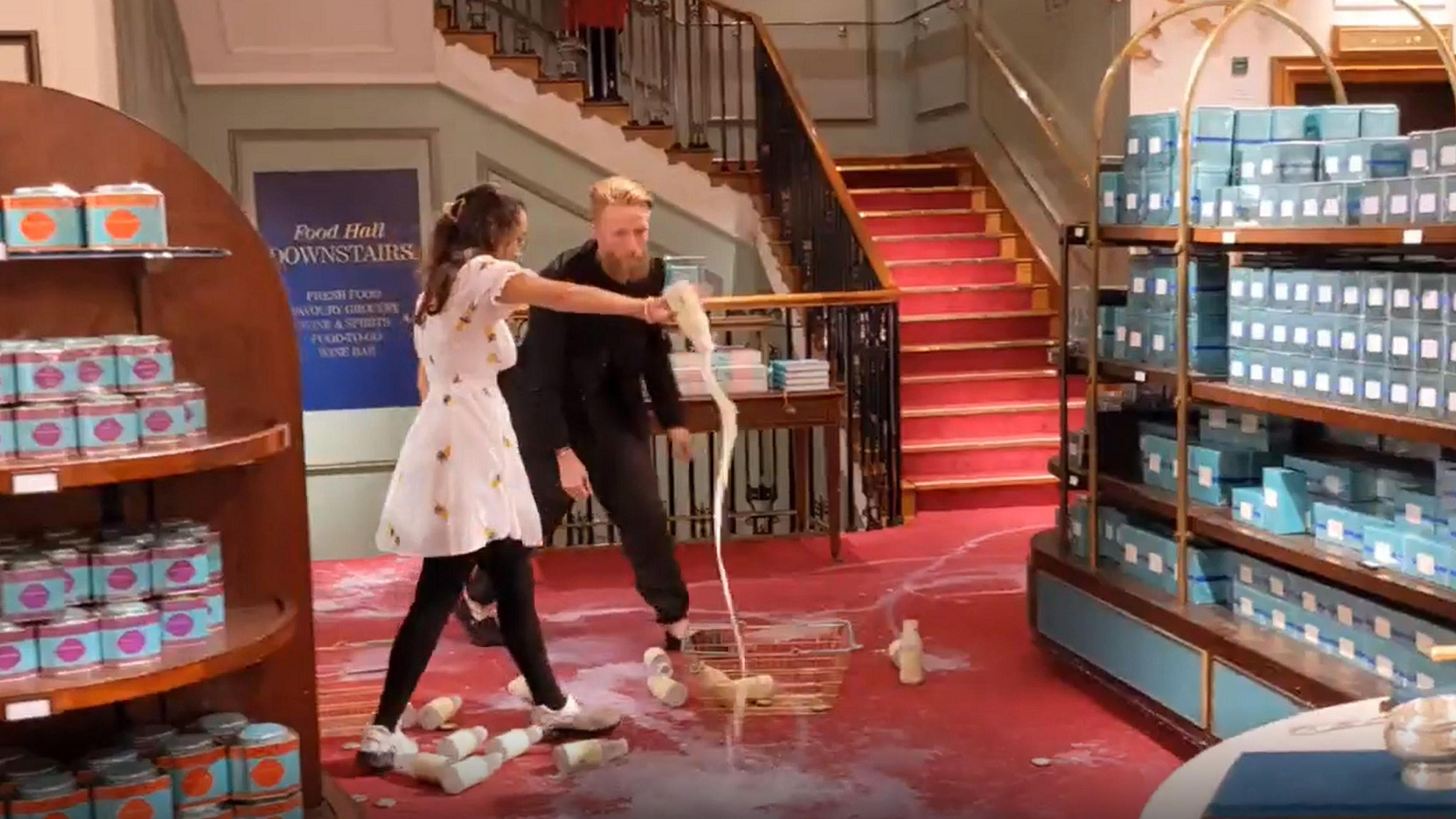 Grab from video issued by Animal Rebellion of supporters of Animal Rebellion - a man wearing black with a big beard and a lady wearing a white dress with brown hair - pouring out milk in Fortnum & Mason in London.