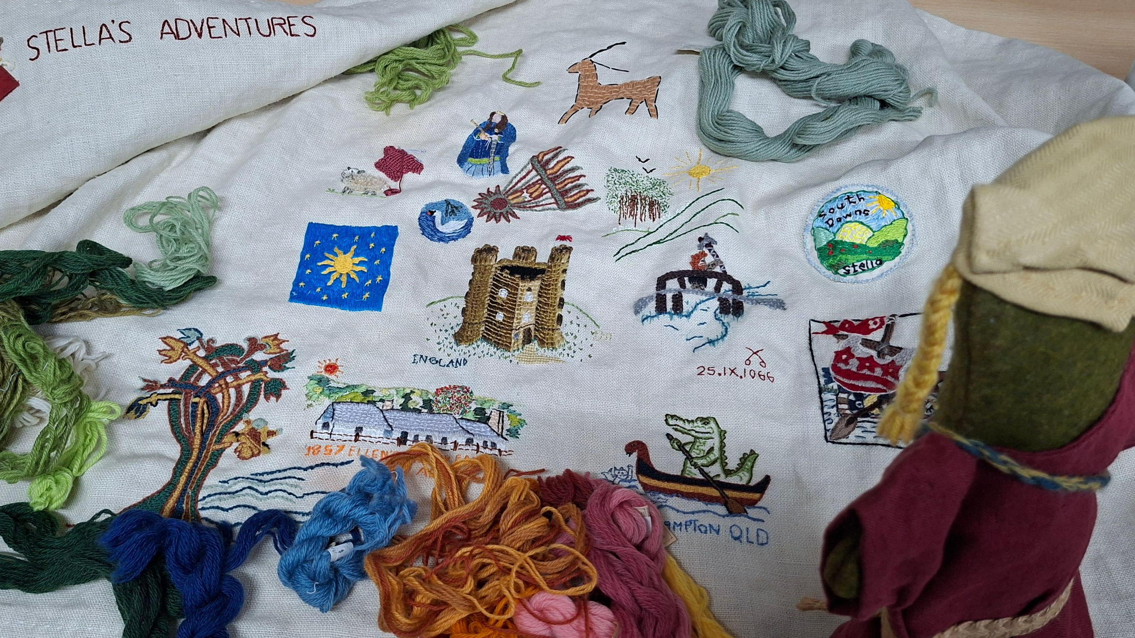 A white one metre square piece of linen with 14 images sewn onto it, such as trees, a crocodile in a rowing boat, flowers and animals.