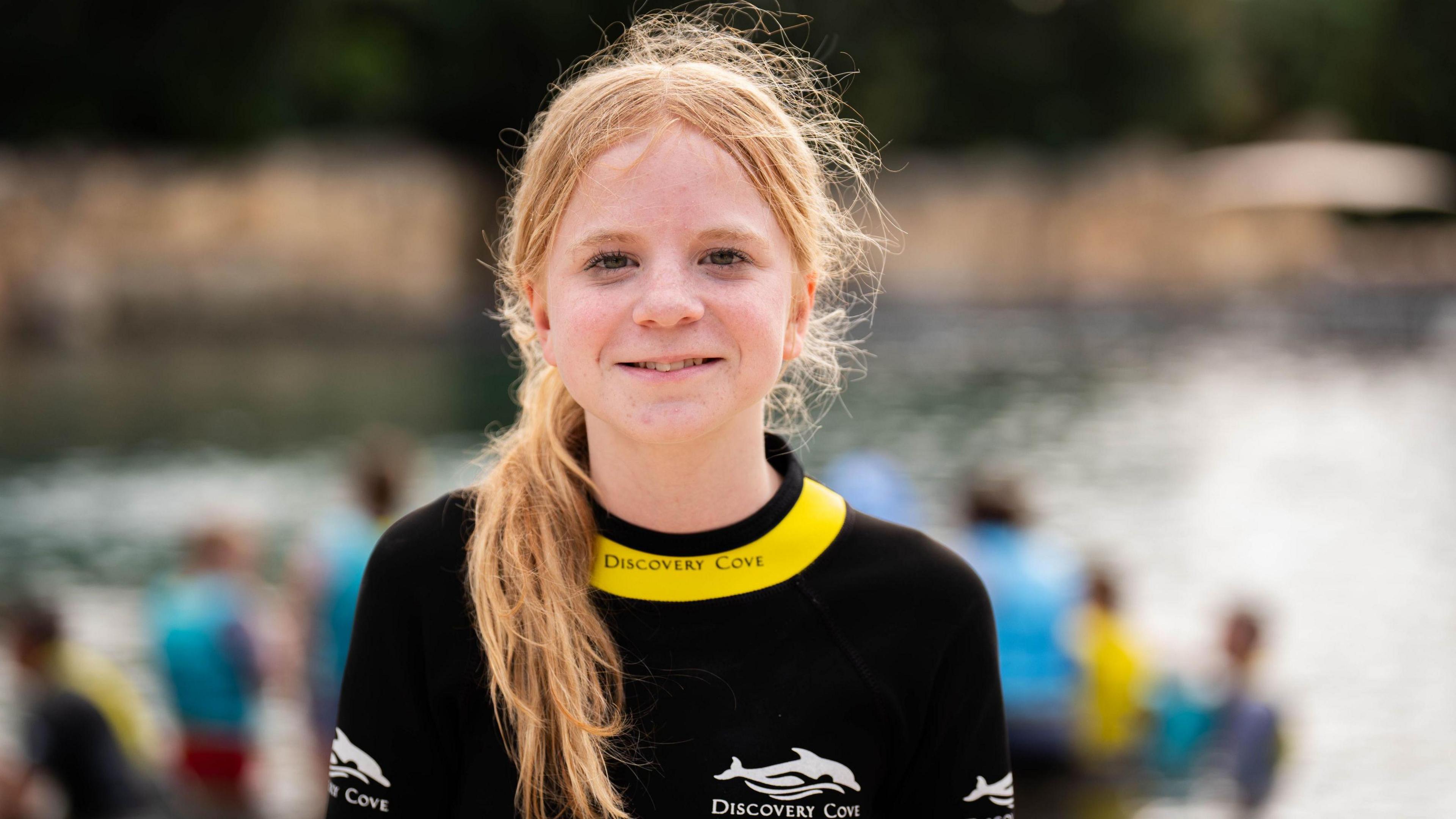 Lottie has strawberry blonde hair that is tied on a ponytail. She is wearing a black and yellow wetsuit that says "discovery cove"