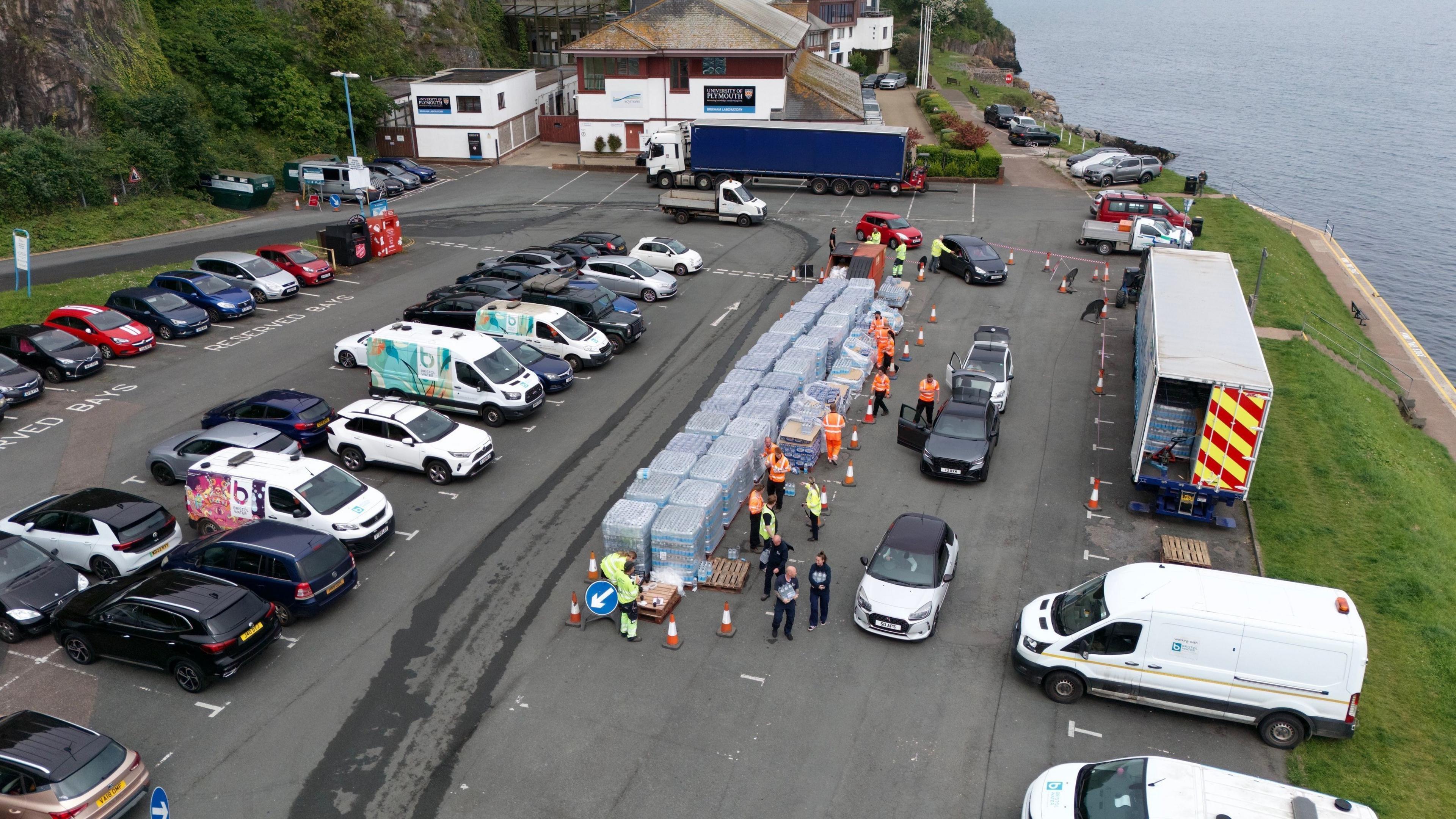 A shot from above a car park with pallets of water bottles stacked in the middle and cars queueing to collect the bottles.  There are people in high-vis jackets handing out the packs of water bottles. 