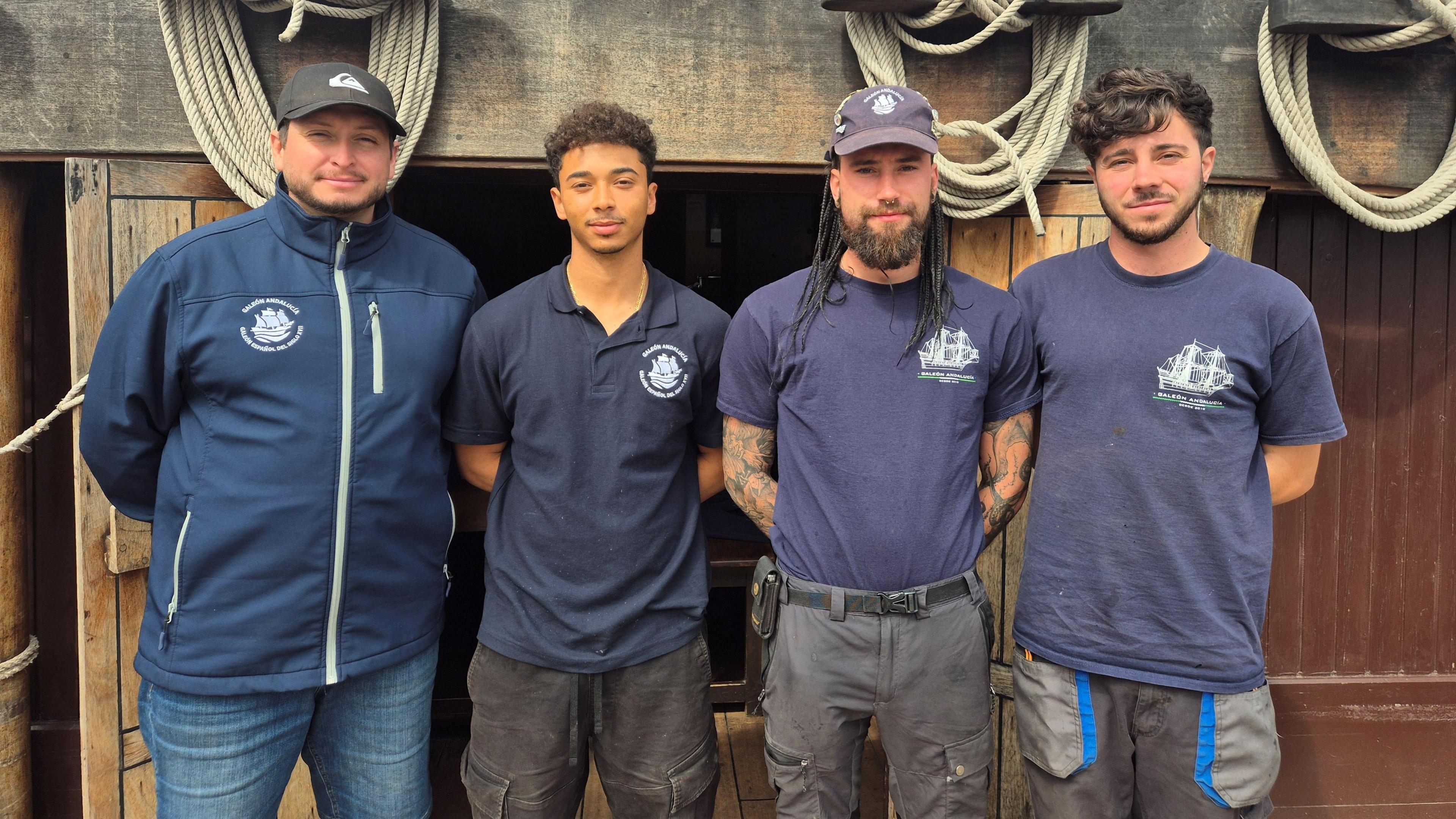 Four members of the Andalucia's crew, all wearing dark blue jackets or shirts and jeans. Two of the group have baseball caps on. They are standing on the deck of the ship.