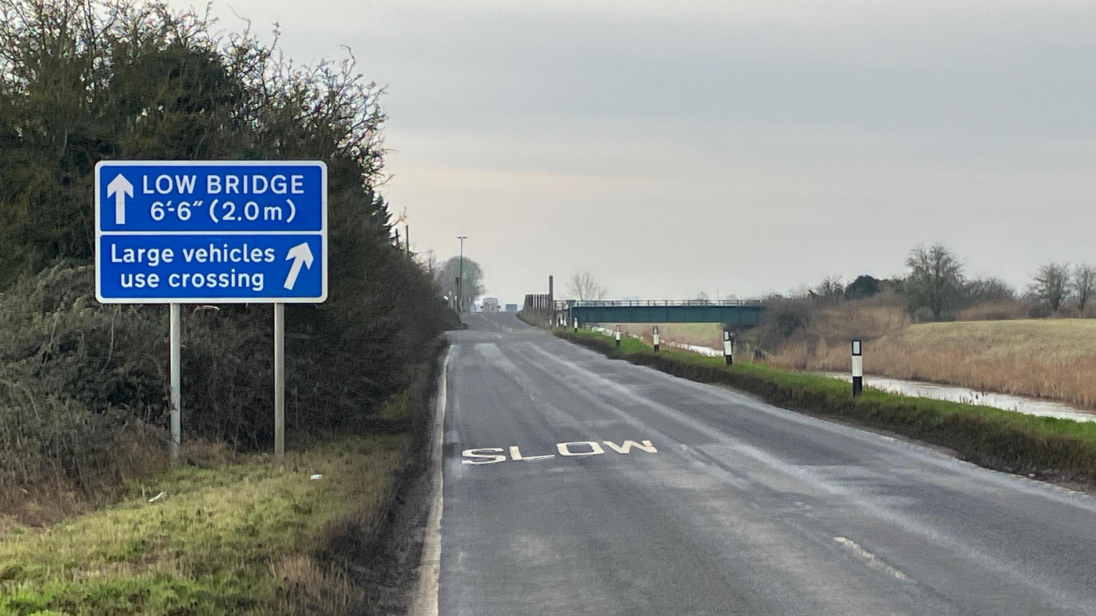 A large blue traffic sign on the approach to a fork in the road that takes vehicles under a 2m (6 foot 6 inch) underpass or alternatively over a manually operated level crossing gate. The lines on the road are barley visible but the word 'STOP' is clearly seen on the left hand side. The Sixteen Foot Drain runs parallel with the road.