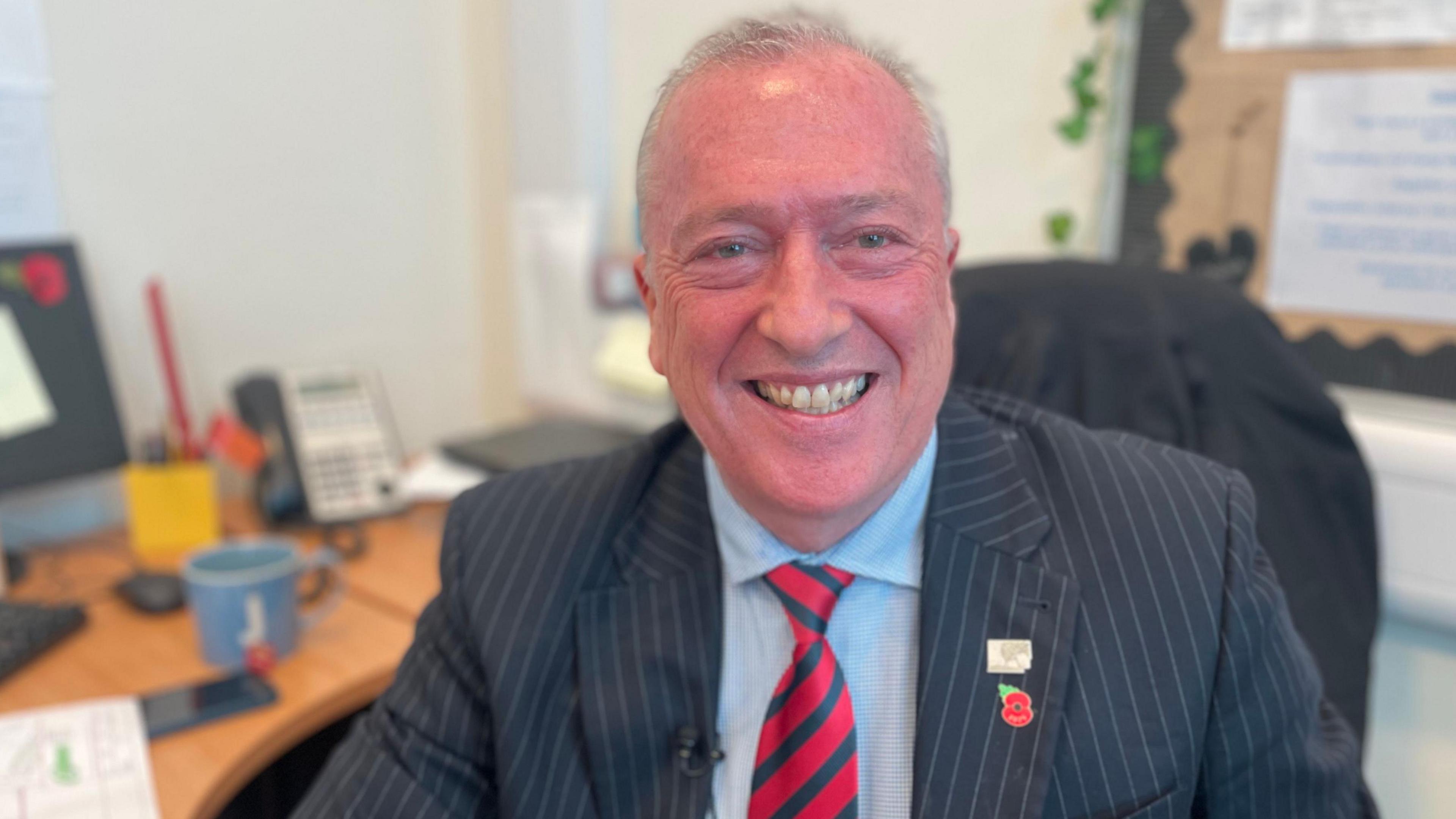 A man with short grey hair in a pinstripe suit jacket and a red and dark green striped tie. He is smiling and sitting at his desk in an office with a pinboard behind him.
