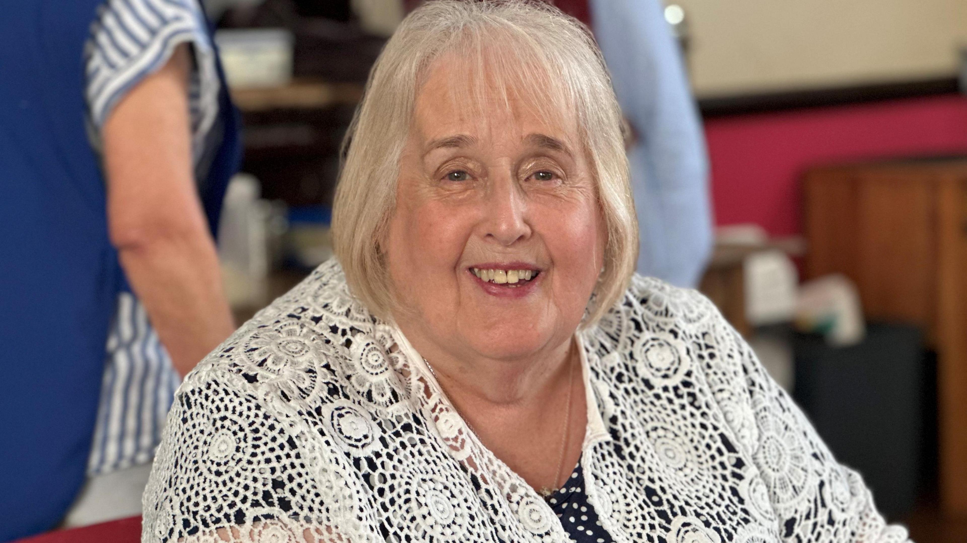 Jackie Hughes, wearing a white patterned crocheted top over a black blouse. She has grey hair cut in a bob.